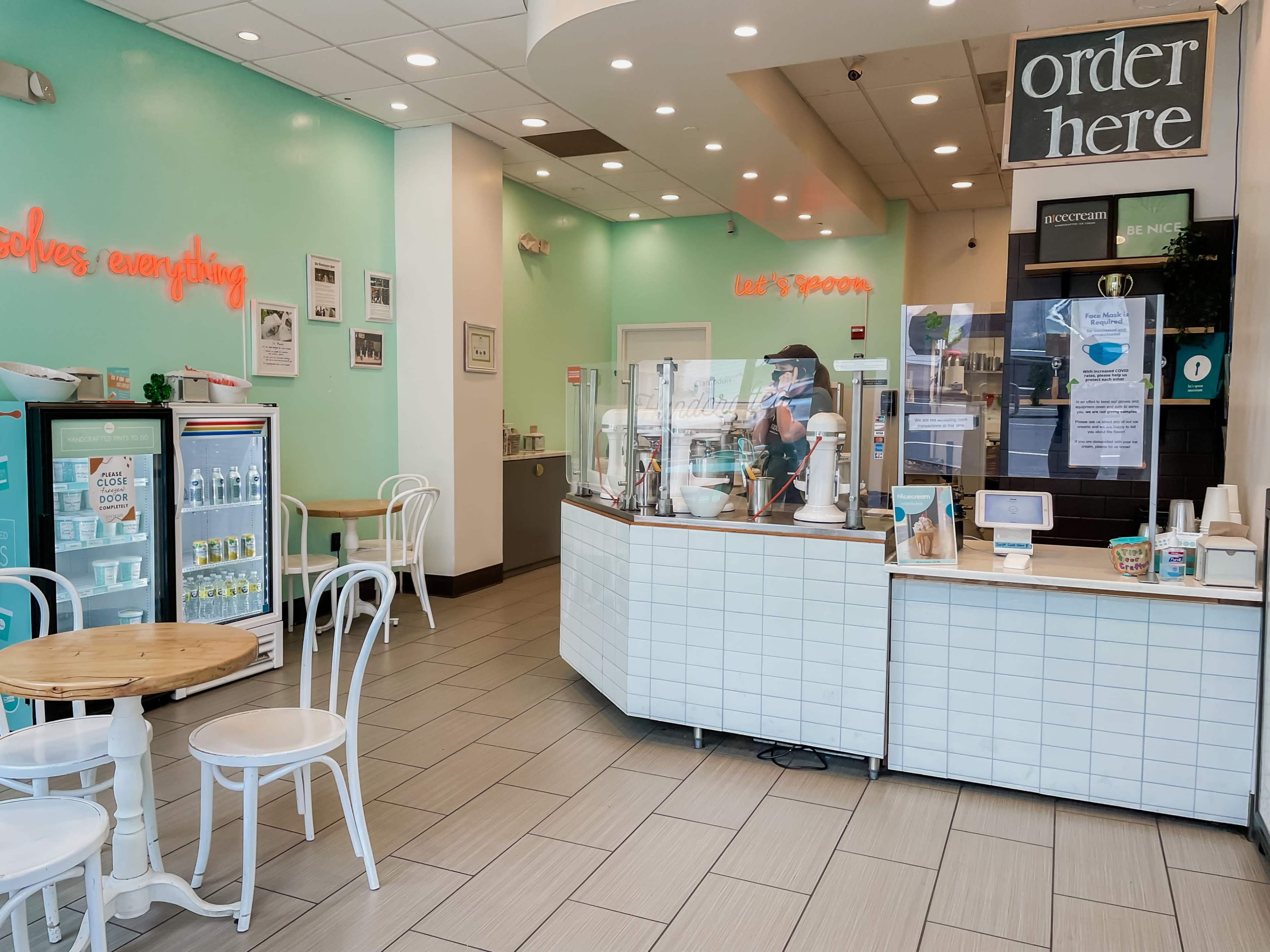 The image shows a modern café interior with mint-green walls, a counter for ordering, a seating area with white chairs and tables, and a display case for food and beverages.