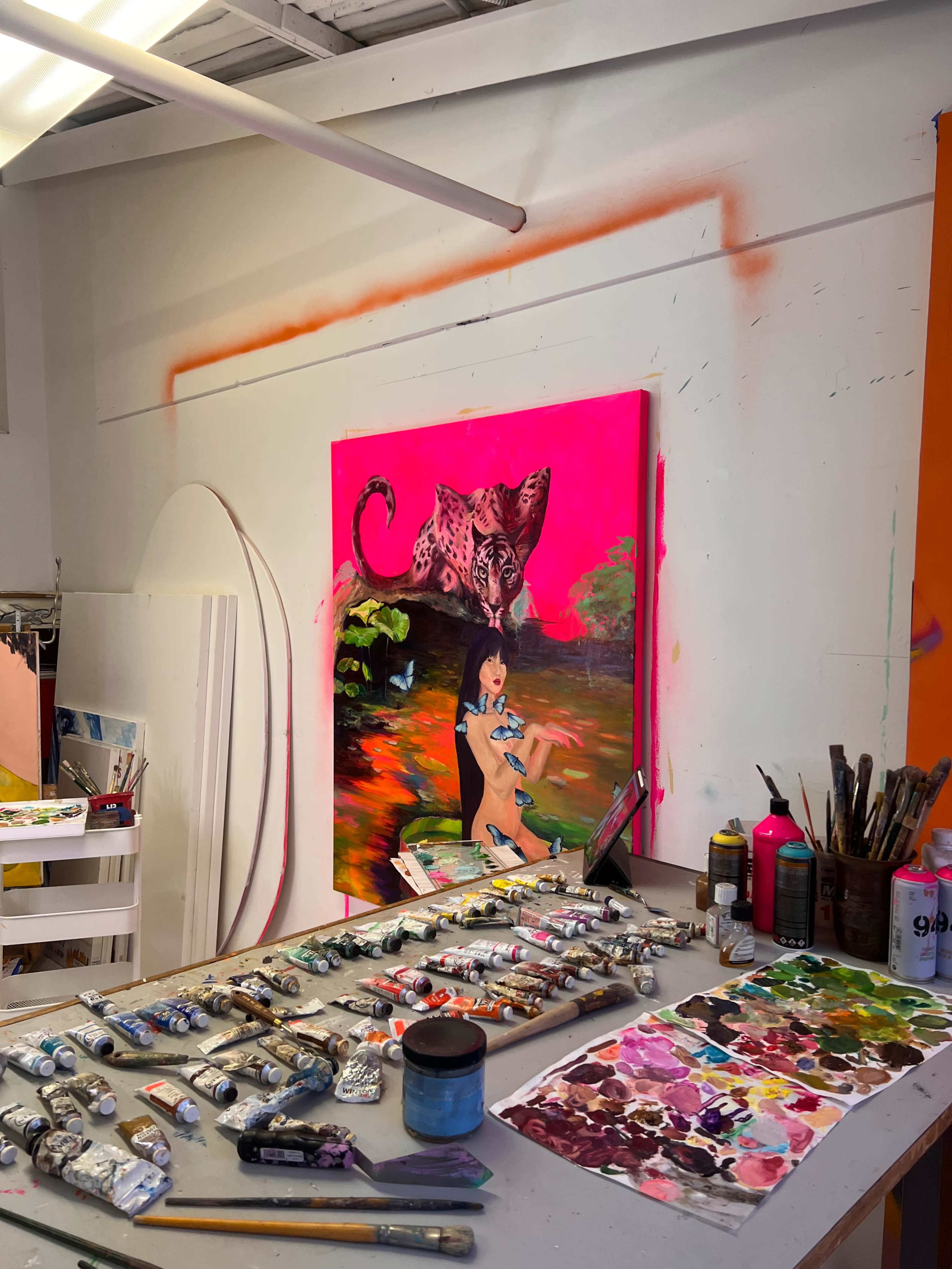 An art studio features a vibrant painting of a girl and a leopard on a bright pink canvas, with various paint tubes and brushes arranged on a worktable in the foreground.