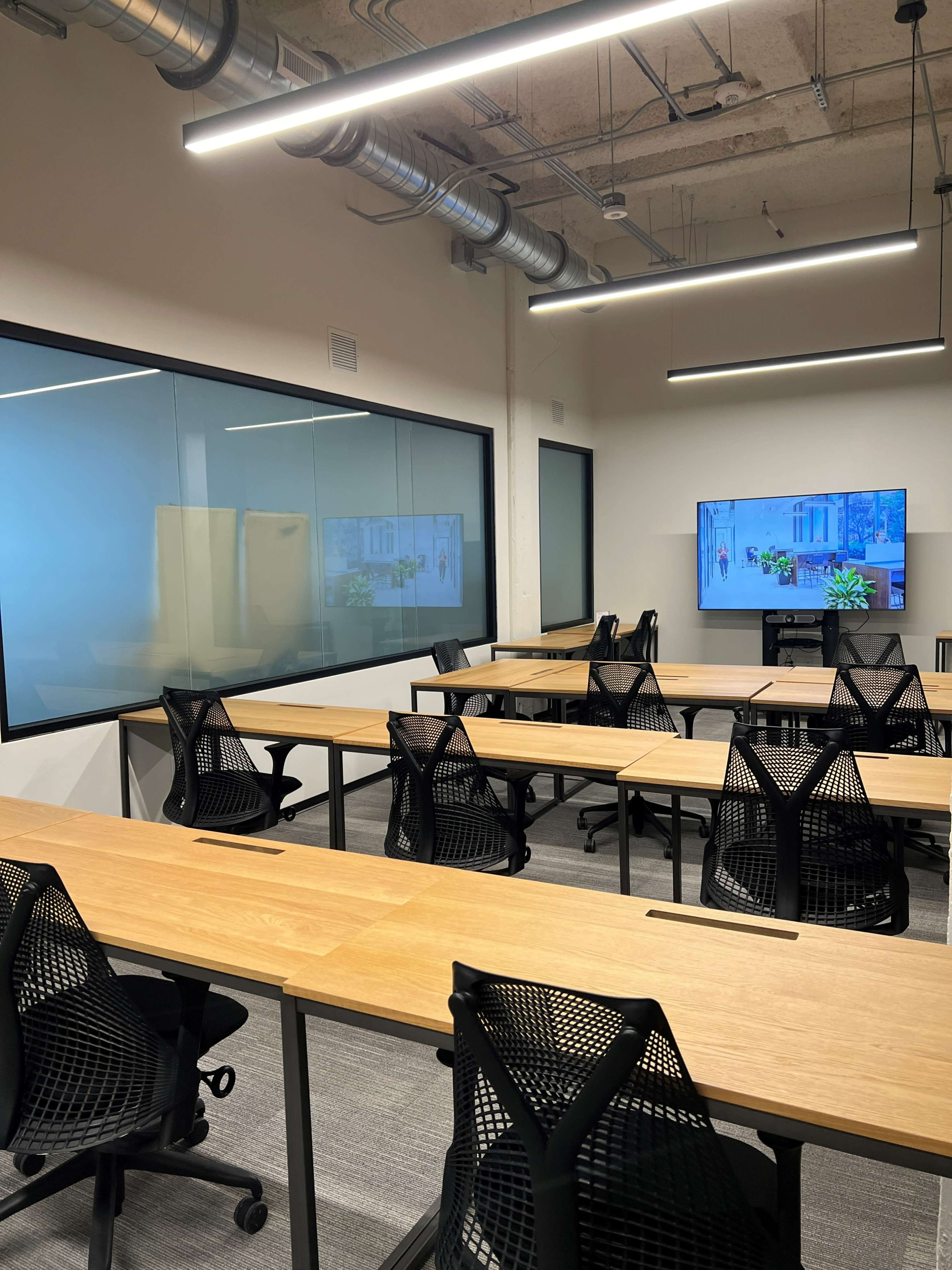 A modern conference room featuring several wooden tables arranged for a meeting, surrounded by black mesh chairs and a large screen on the wall.