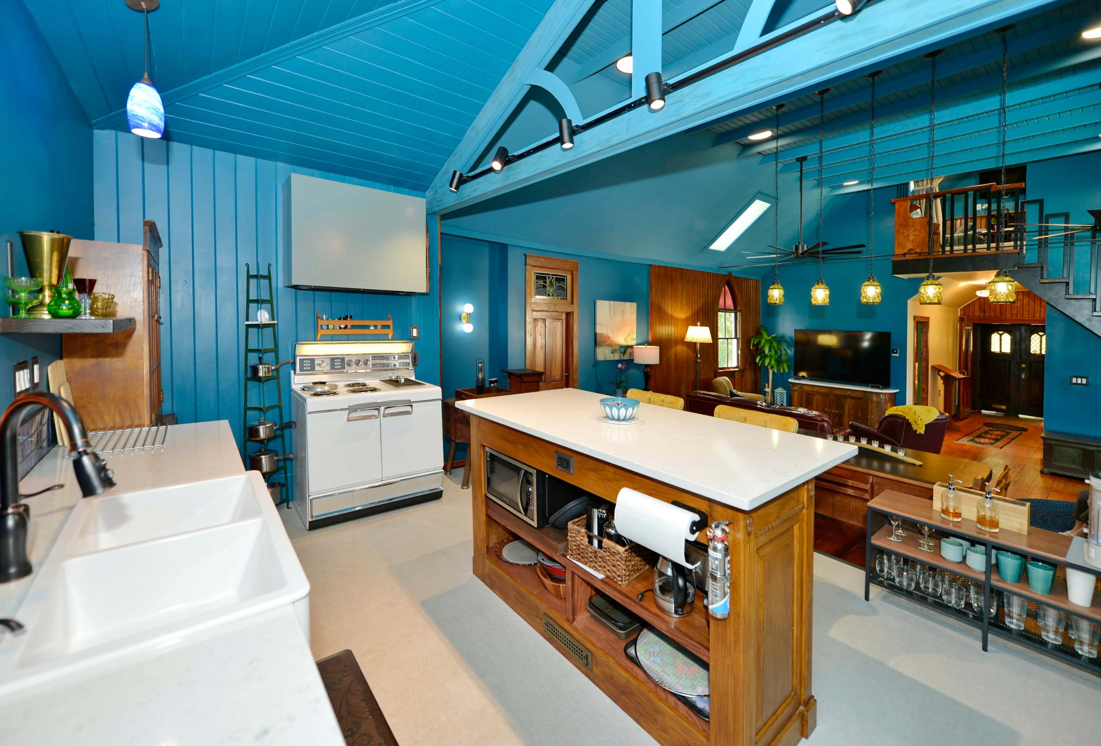 The image shows a brightly colored kitchen with blue walls, featuring a central island, a stove, and open space leading to a living area with a staircase.