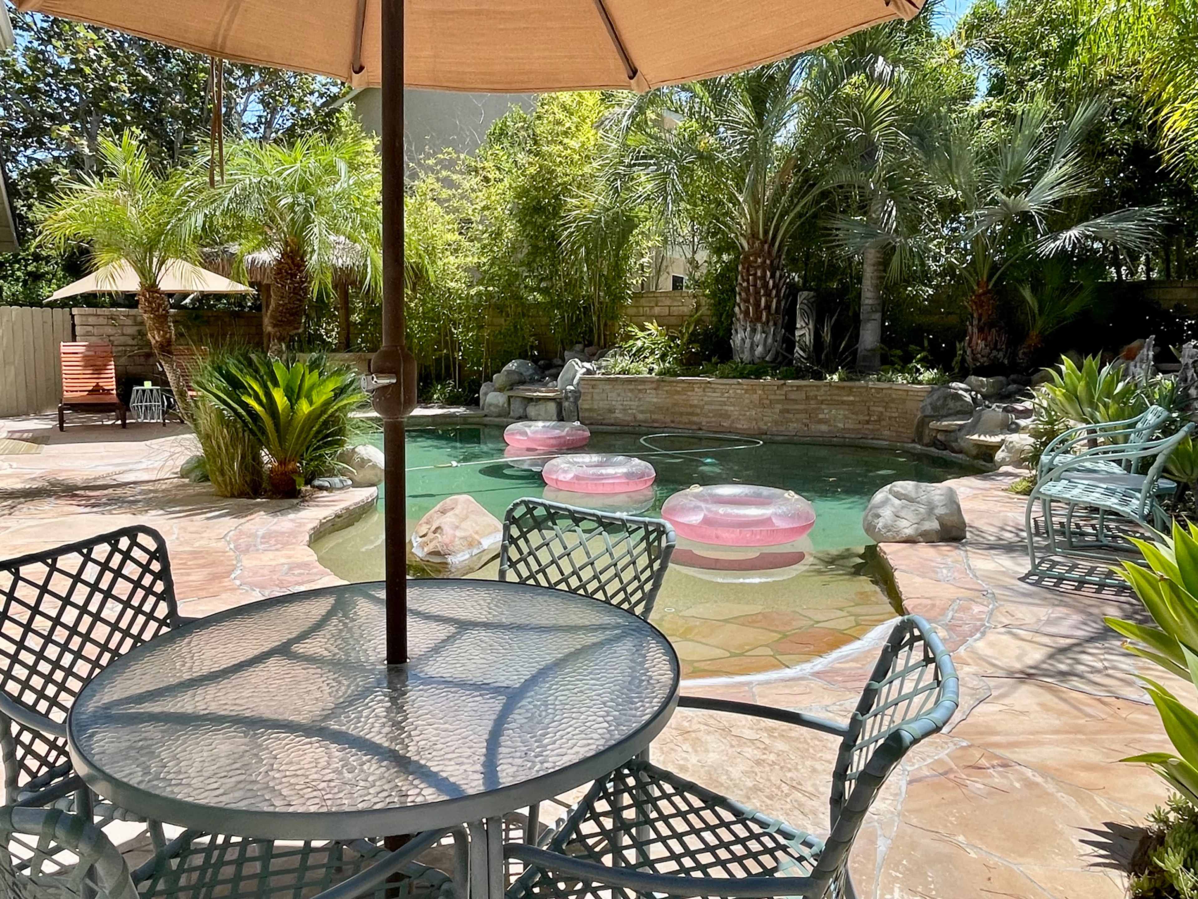 A patio with a table and chairs overlooks a swimming pool surrounded by palm trees and decorative rocks, featuring pink inflatable floats.