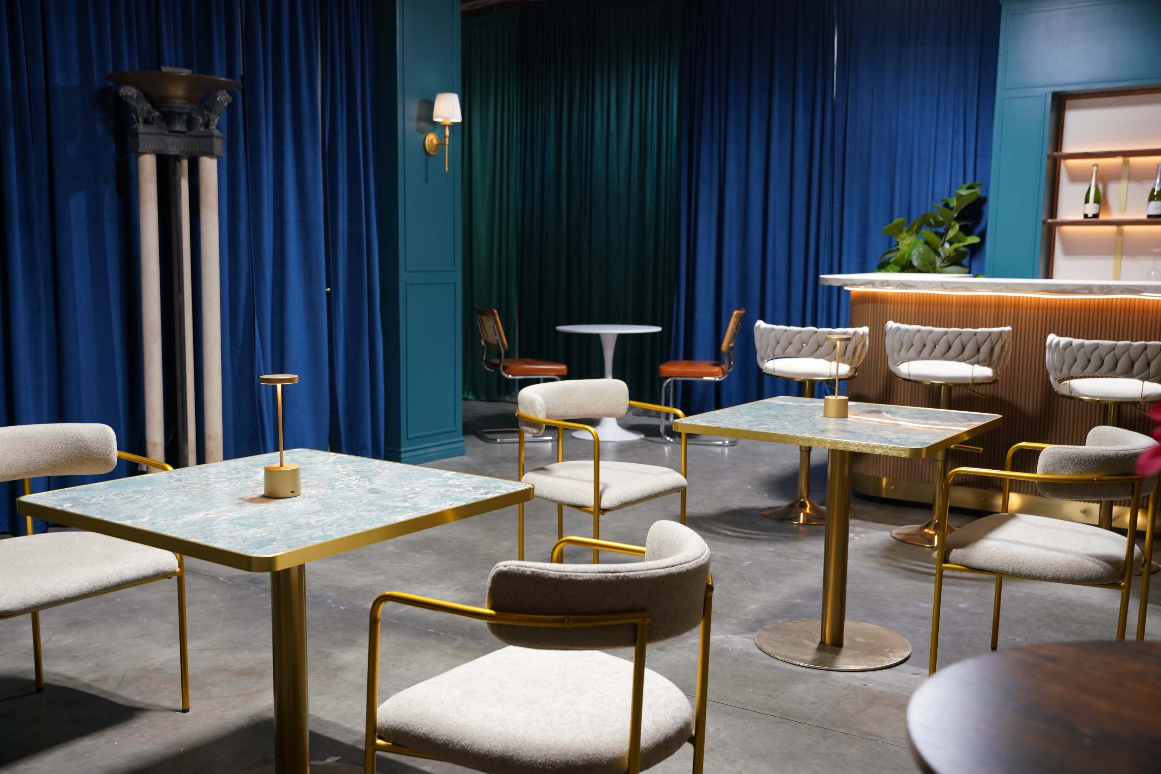 The image shows a stylish indoor space featuring modern tables and chairs, a bar area, and blue curtains.