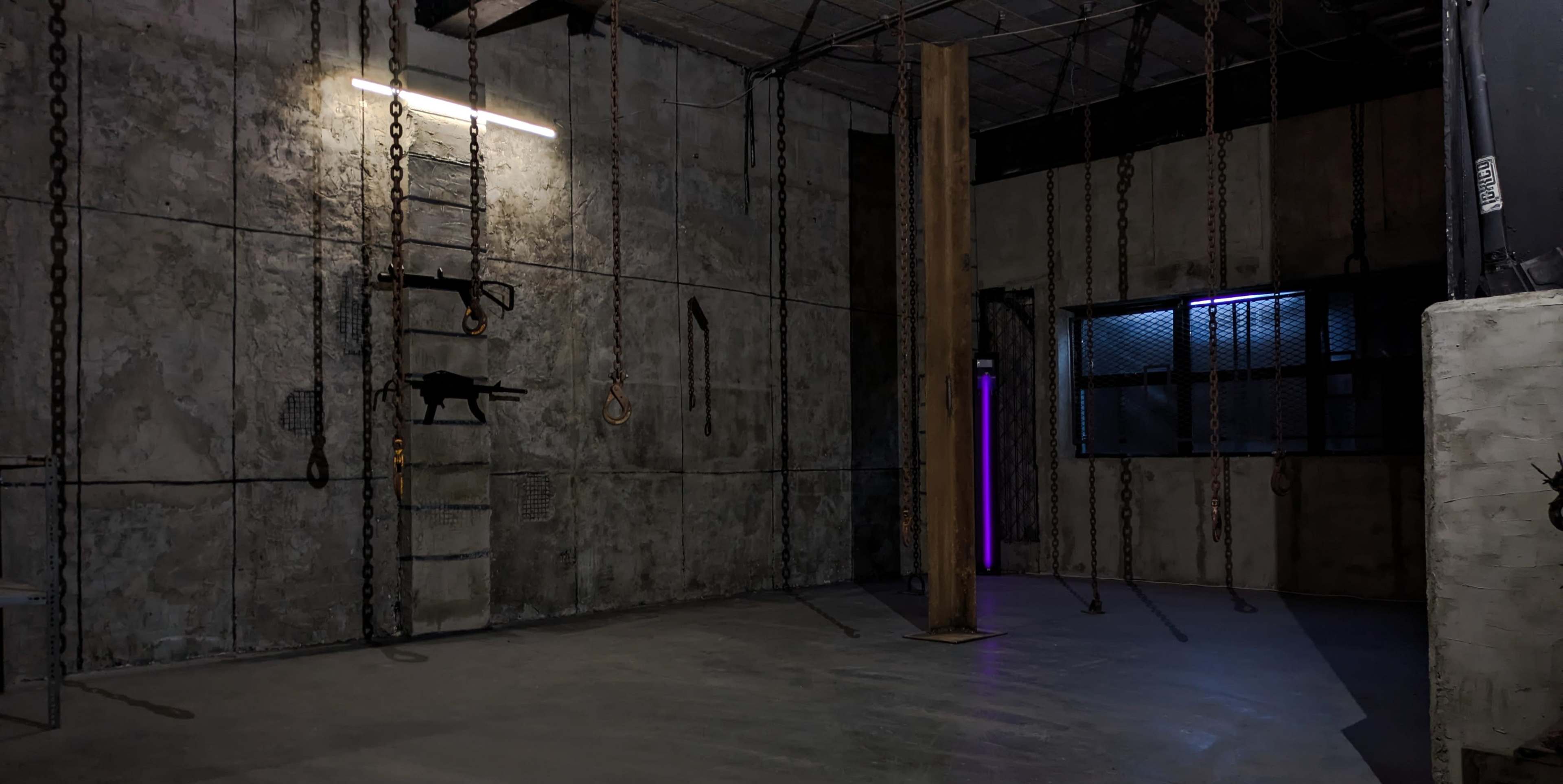 The image shows a dimly lit industrial space with concrete walls, a wooden support beam, chains hanging from the ceiling, a ladder against the wall, and a window with purple lighting.