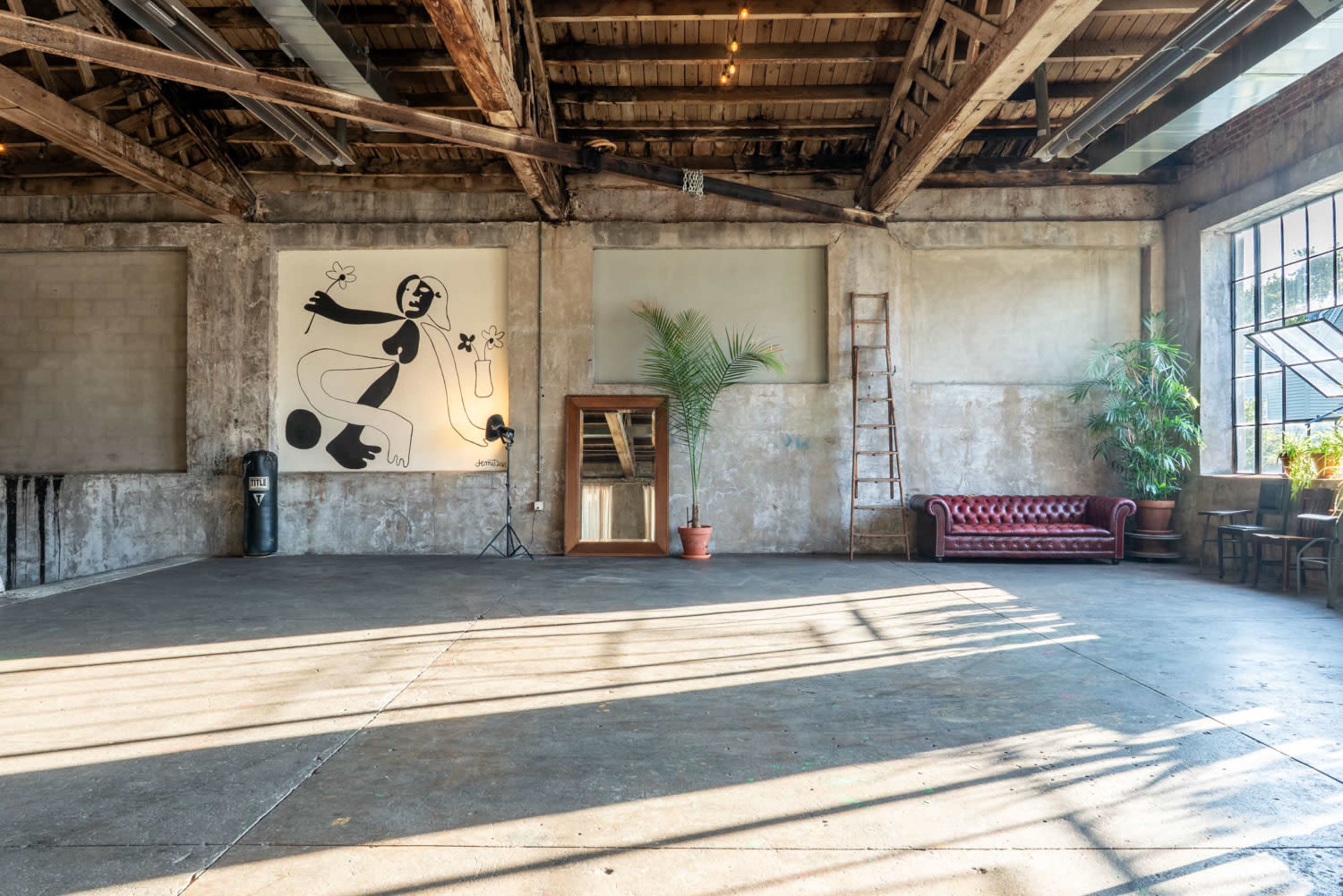 The image shows a spacious industrial interior with exposed wooden beams, a large wall mural, a ladder, plants, and a red couch.