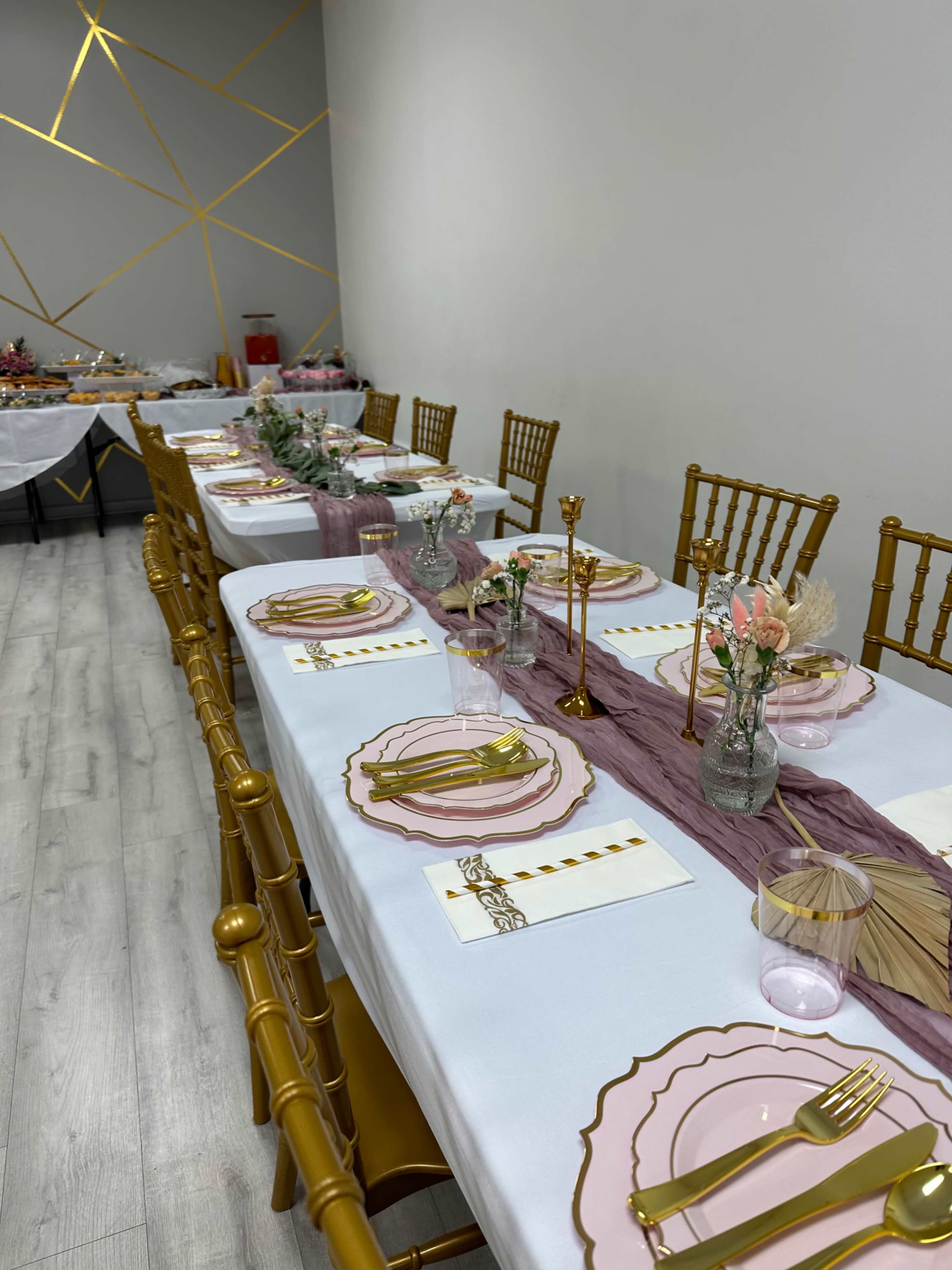 A long dining table is set with pink plates, gold utensils, and small vases of flowers, arranged neatly in a room with gold accent walls.