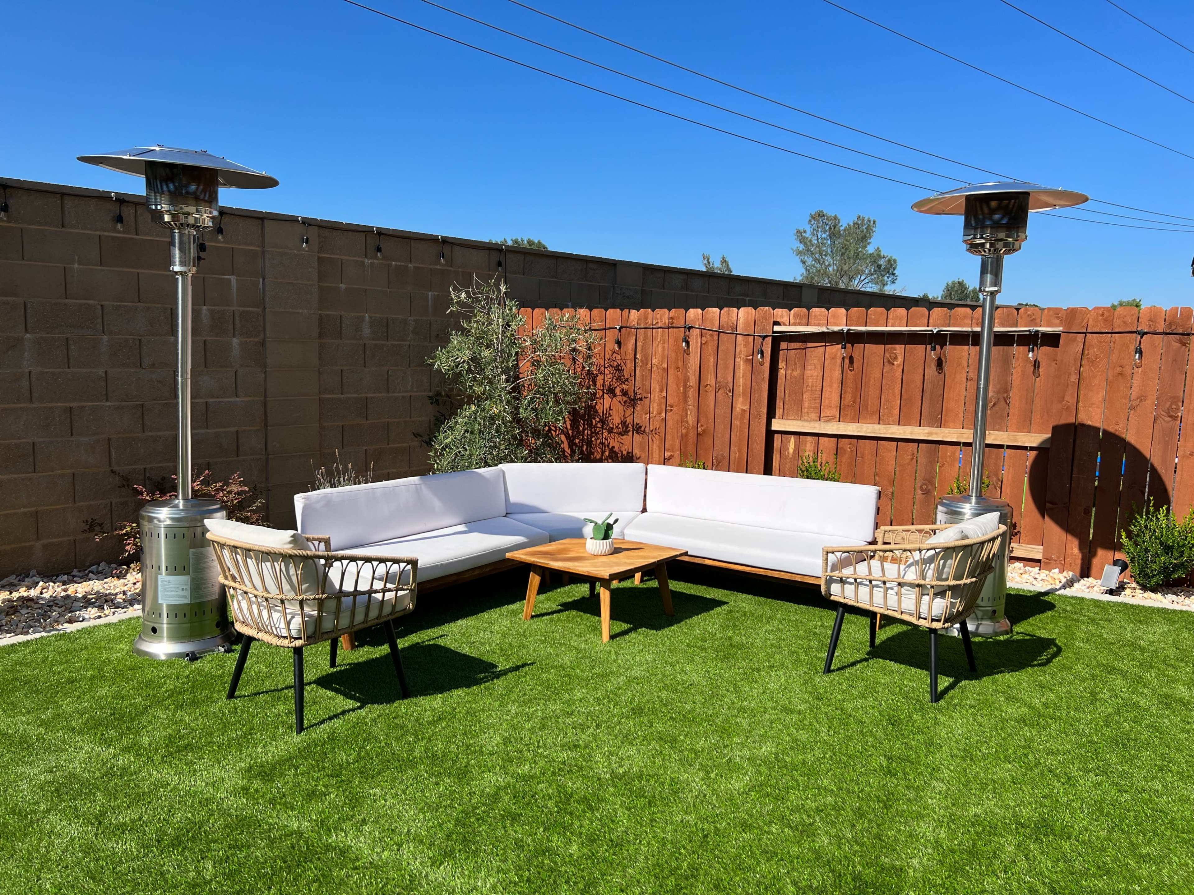 A modern outdoor seating area with a white sectional sofa, two woven armchairs, a wooden coffee table, and gas patio heaters, all set on a green artificial lawn against a wooden fence.