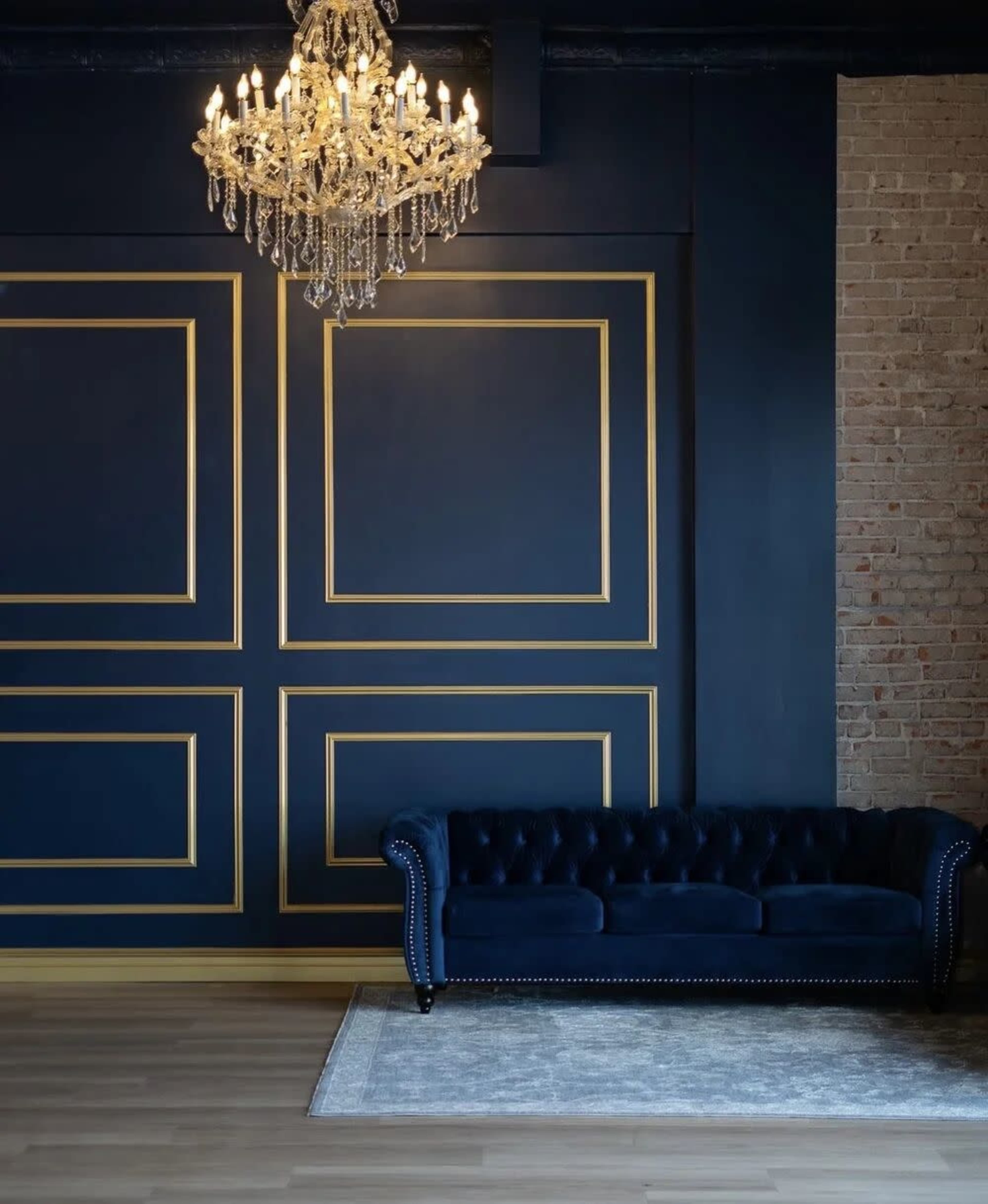 The image features a navy blue wall with gold-framed paneling, a tufted navy blue sofa, a large chandelier, and a light-colored area rug on a wooden floor.