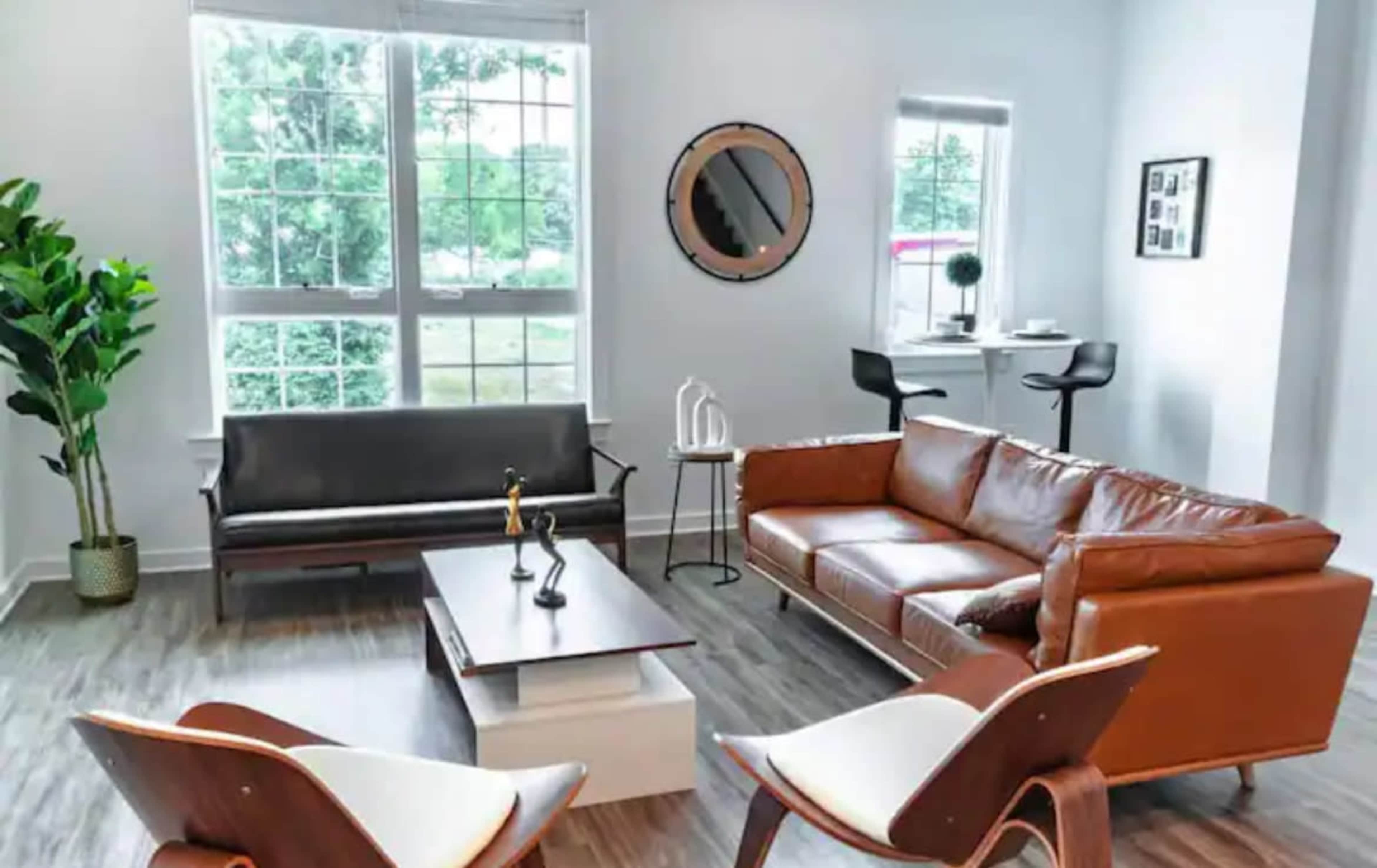 A modern living room with a leather sofa, a wooden coffee table, two armchairs, and large windows letting in natural light.