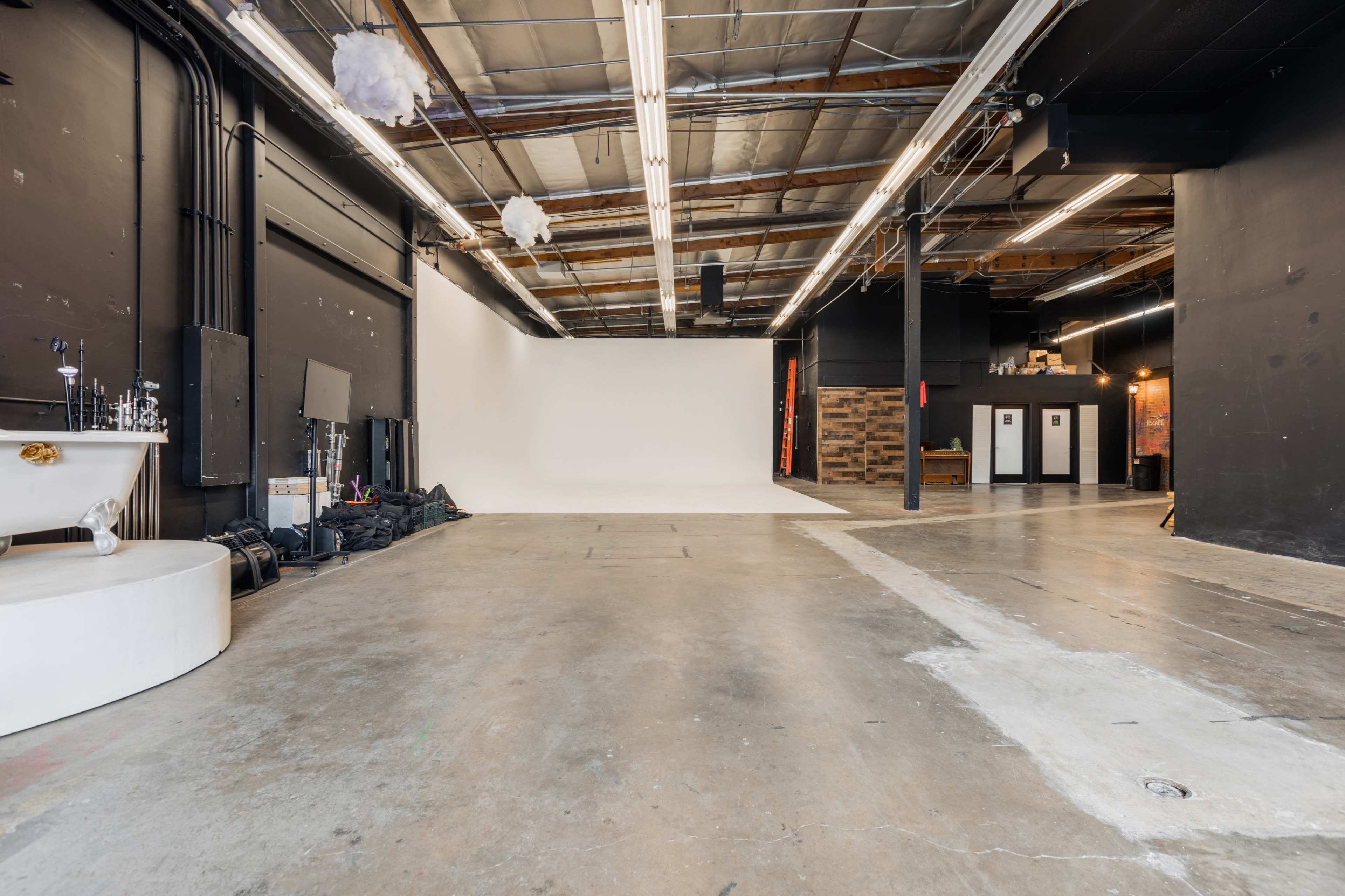 The image shows an industrial-style studio space with a white backdrop, concrete flooring, and various equipment and furnishings along the walls.