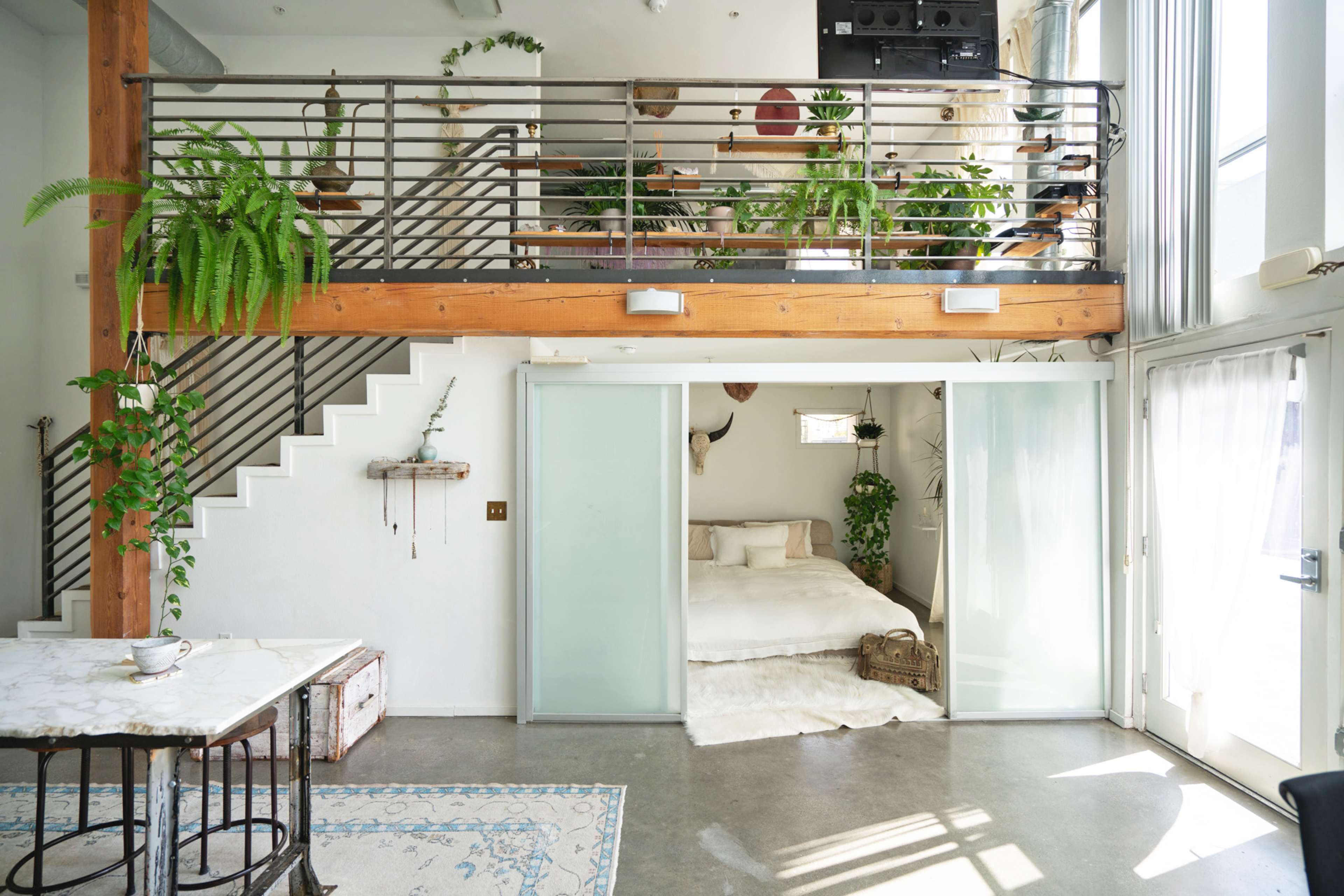 A bright, open living space featuring a dining area, a staircase, a glass sliding door leading to a bedroom, and various plants throughout.