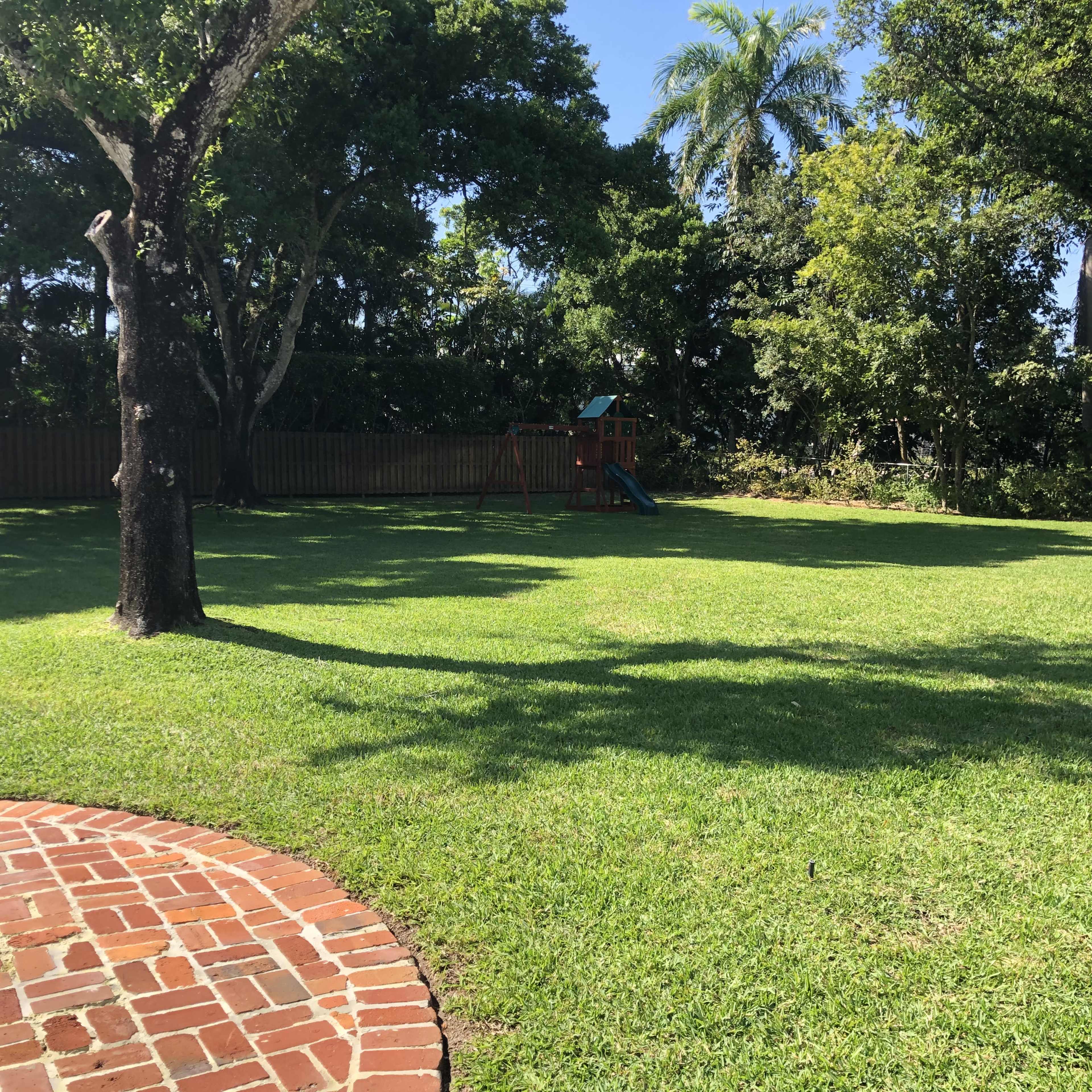 A grassy backyard features a wooden playground set among scattered trees and a paved circular patio.