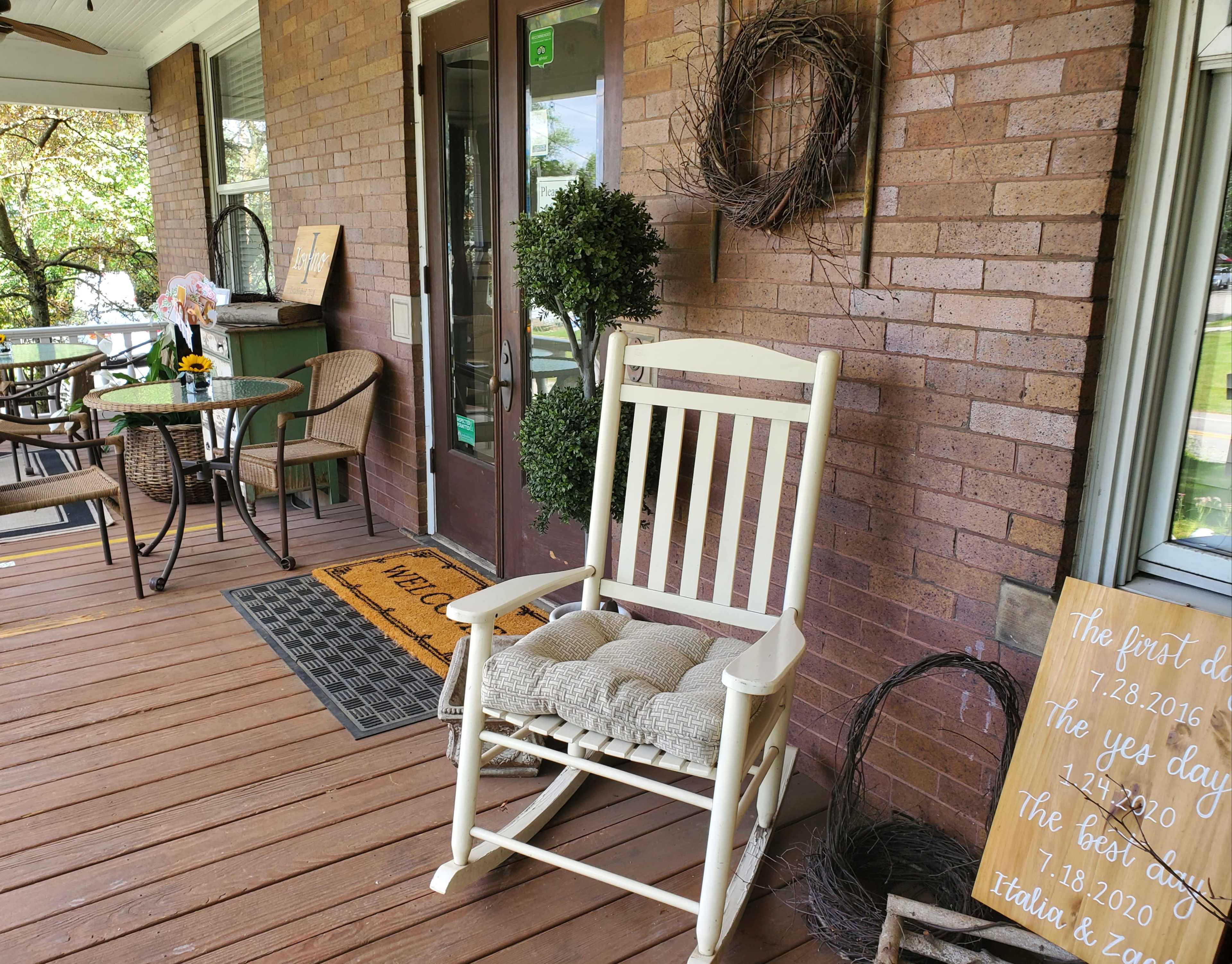 A porch with a white rocking chair, a small table set for dining, and a decorative welcome mat along with wooden signage.