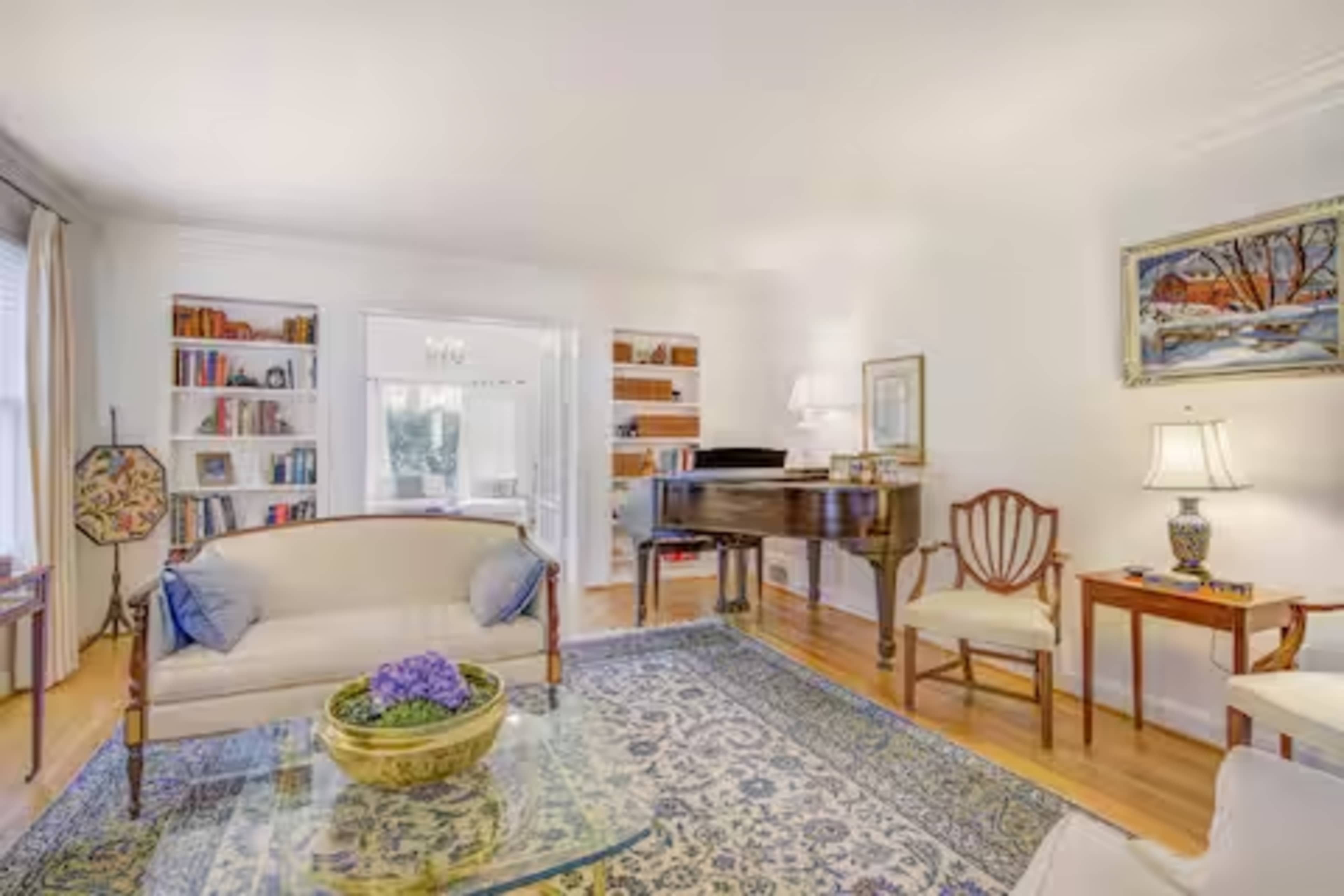 A bright living room features a beige sofa, a wooden piano, bookshelves, and decorative lamps.