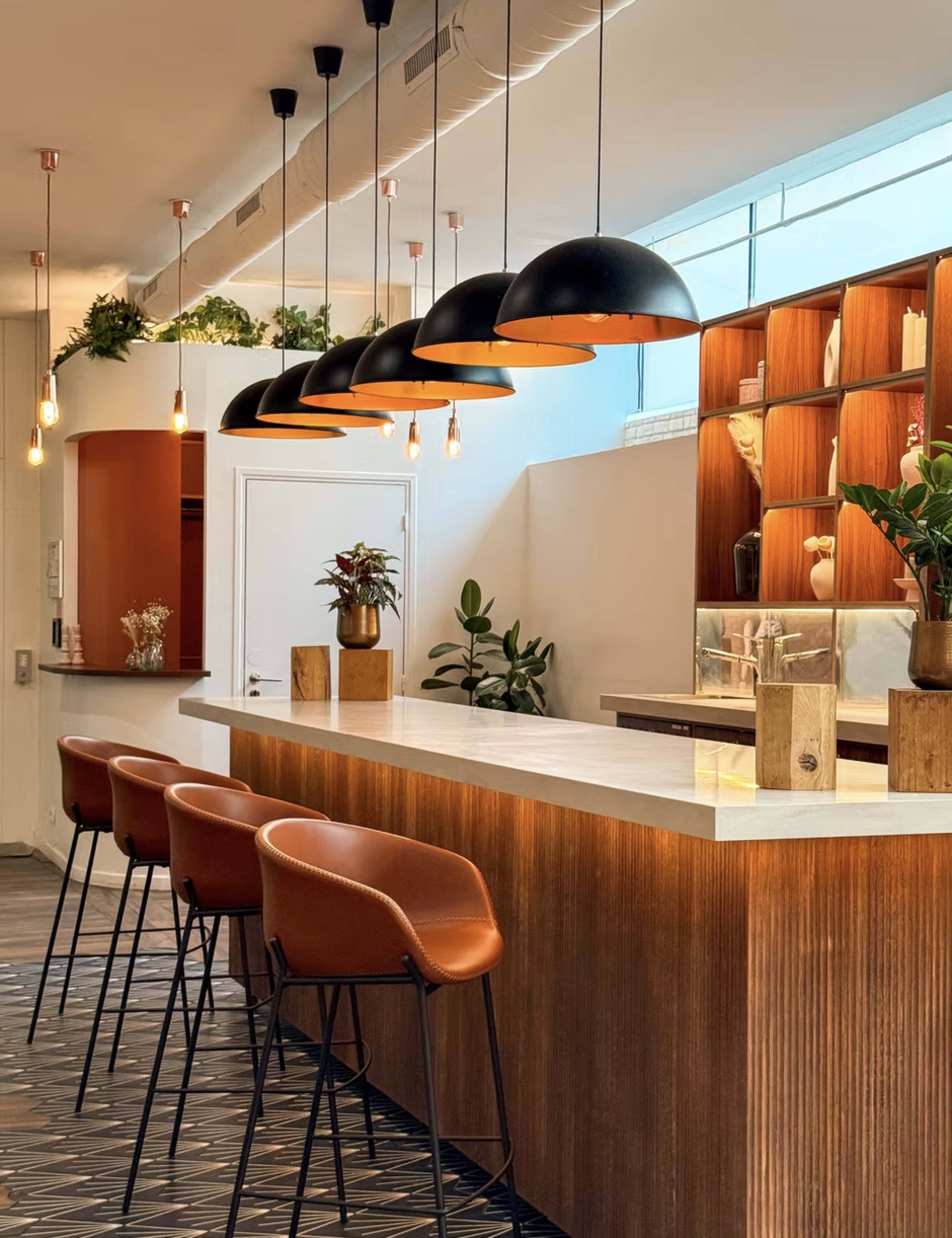 A modern bar features a light-colored countertop, dark pendant lights, and brown seating against a backdrop of wooden shelves and greenery.