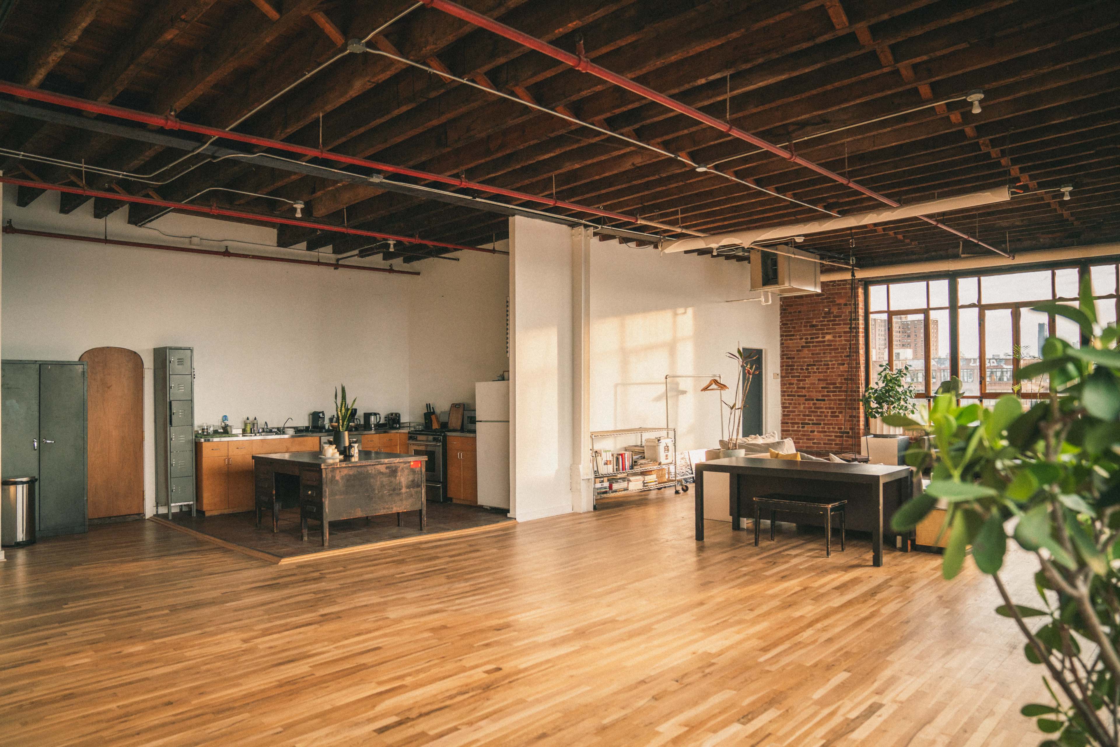The image shows a spacious industrial-style loft with wooden floors, exposed ceiling beams, and a kitchen area to the left.