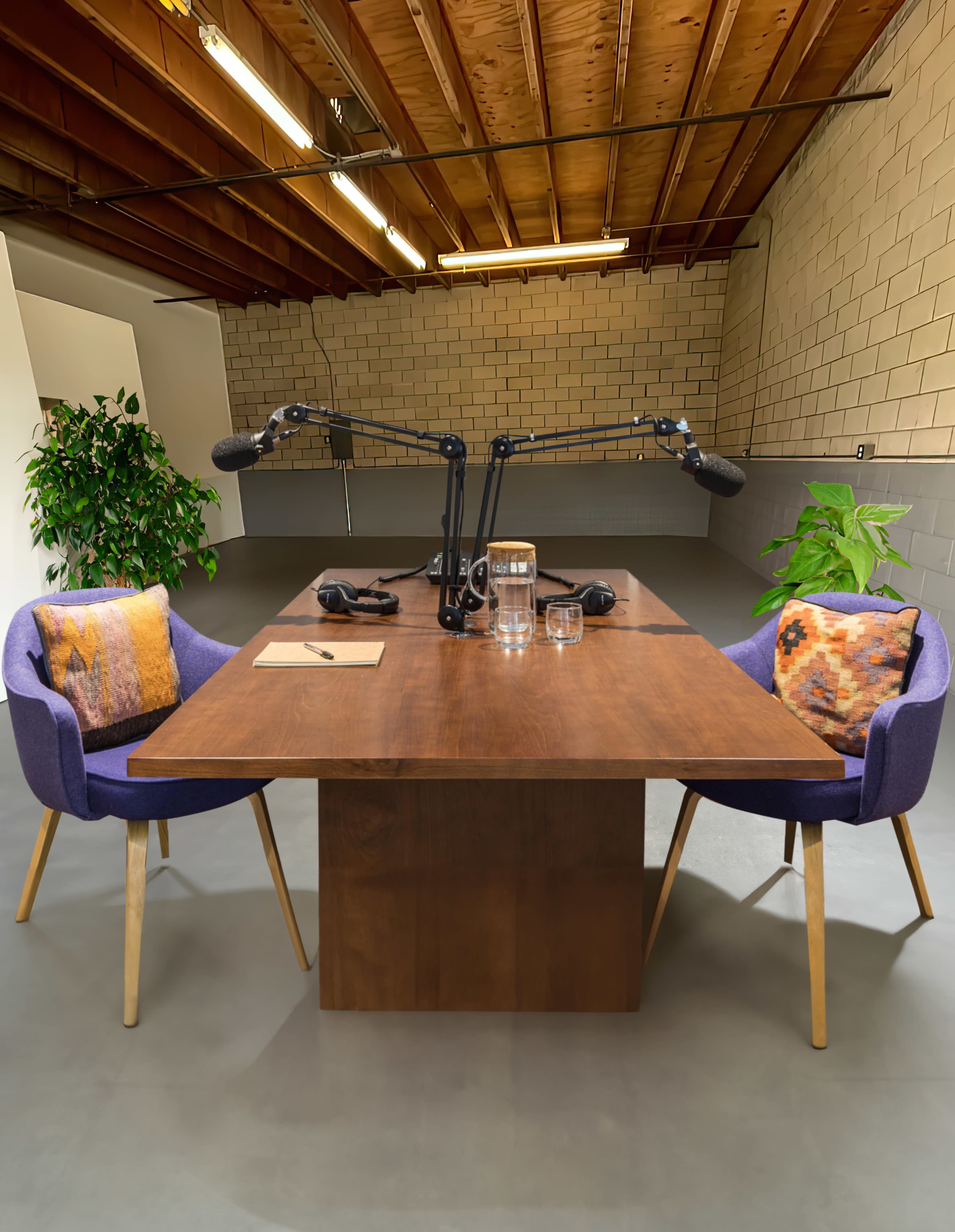 A wooden table is set in a studio space with two chairs, each adorned with colorful pillows, and microphones mounted above.