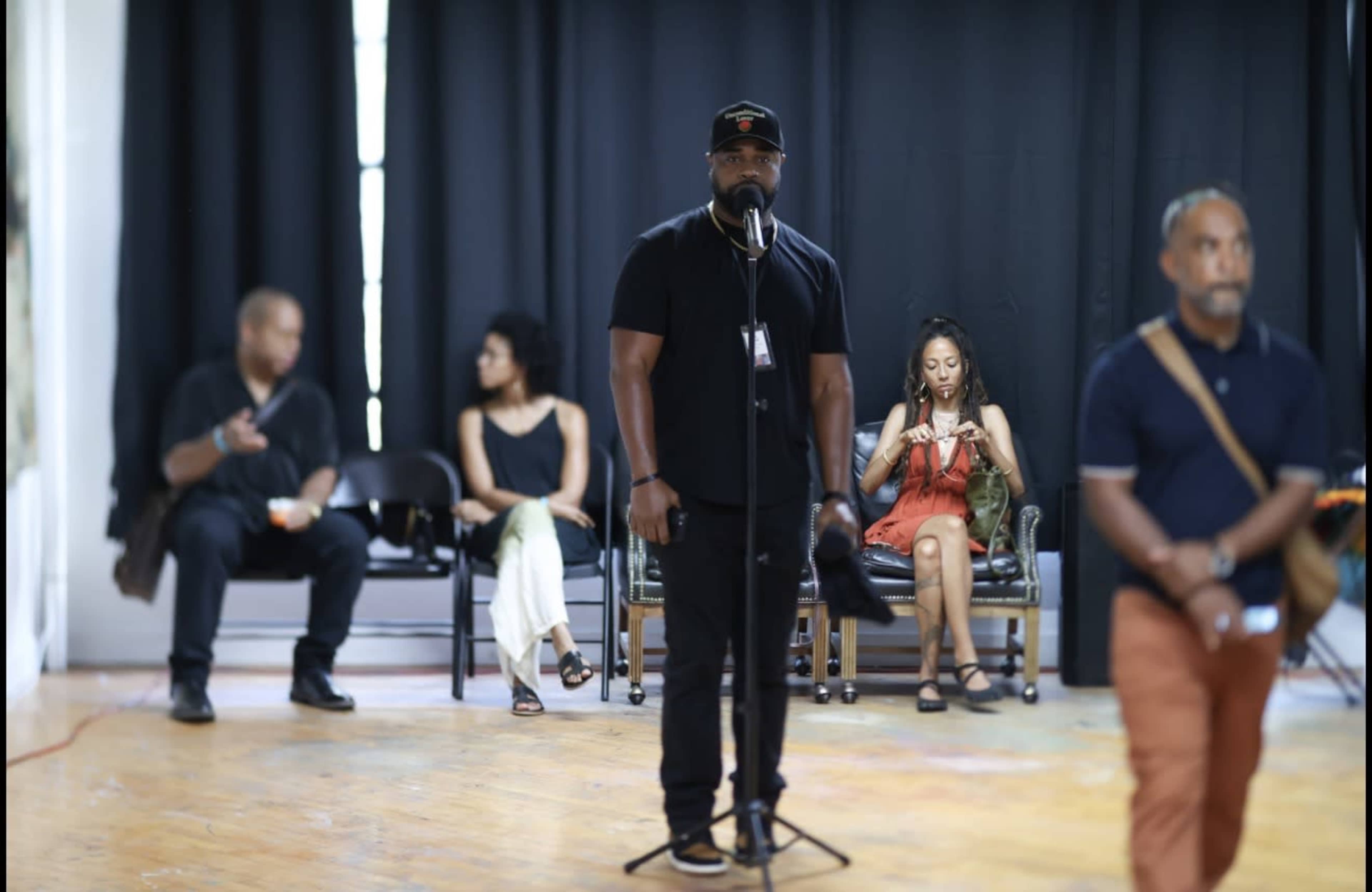 A man stands at a microphone while several individuals are seated in the background, some engaged in conversation and others using their phones.