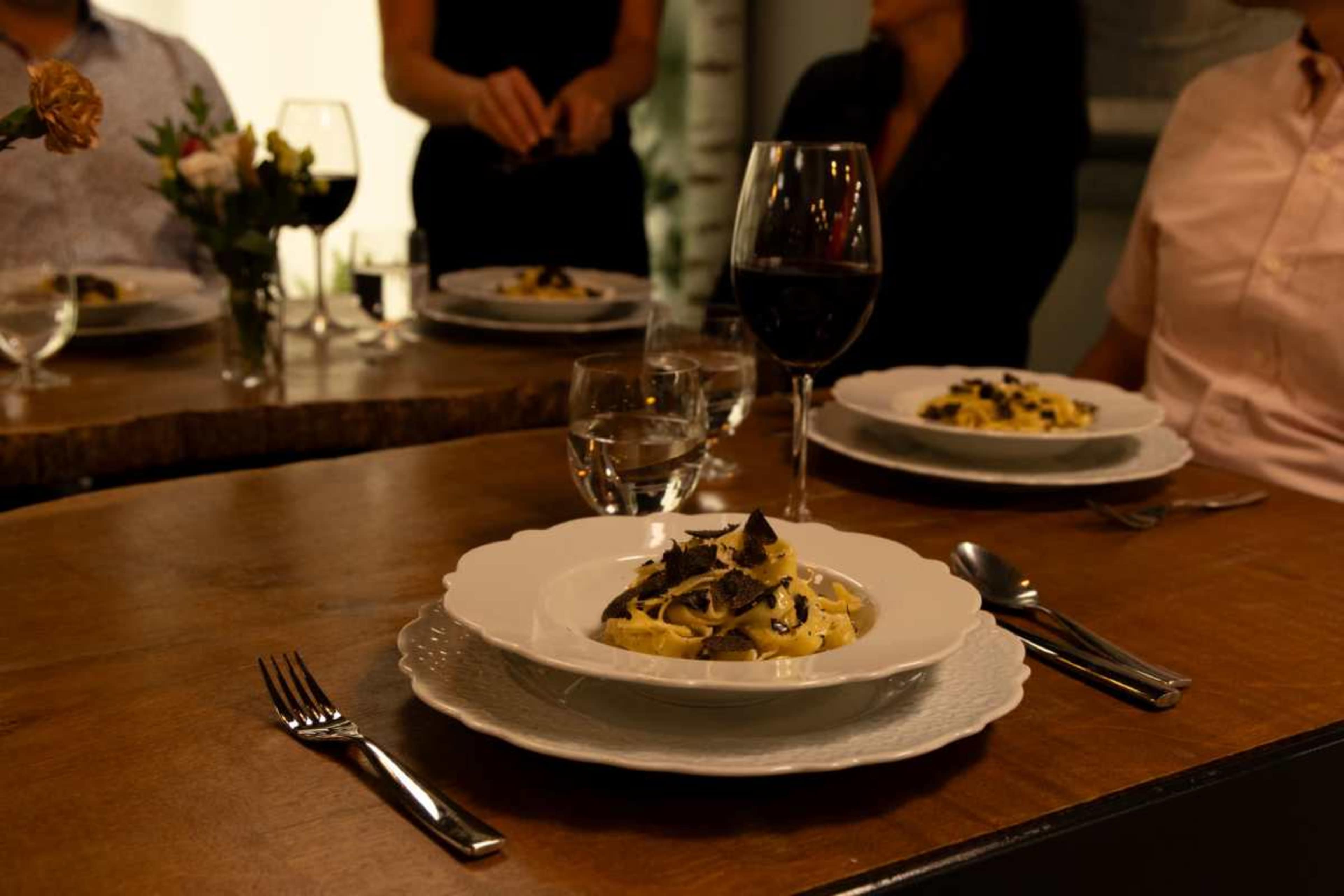 A table is set for a dinner with plates of pasta, wine glasses, and a few diners in the background.