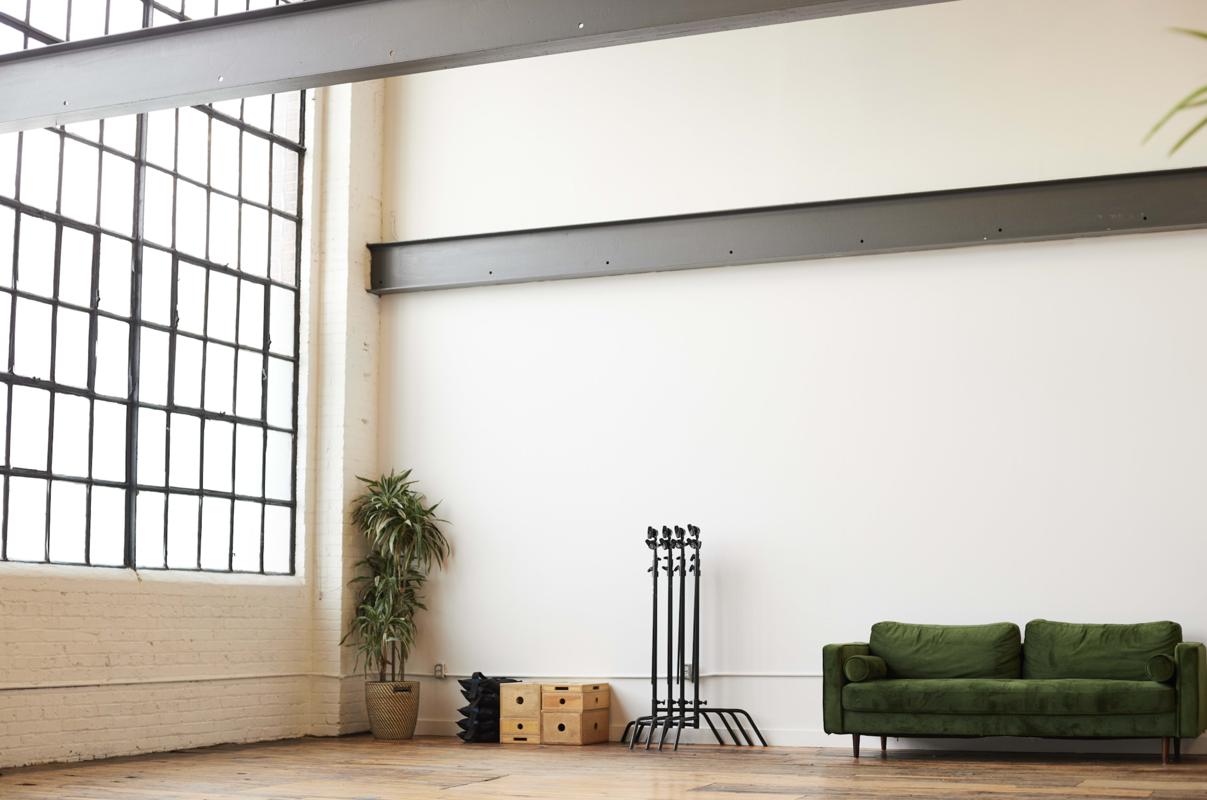 A green sofa sits in a spacious room with large windows, wooden flooring, and minimal decor, featuring a plant and several upright coat racks.