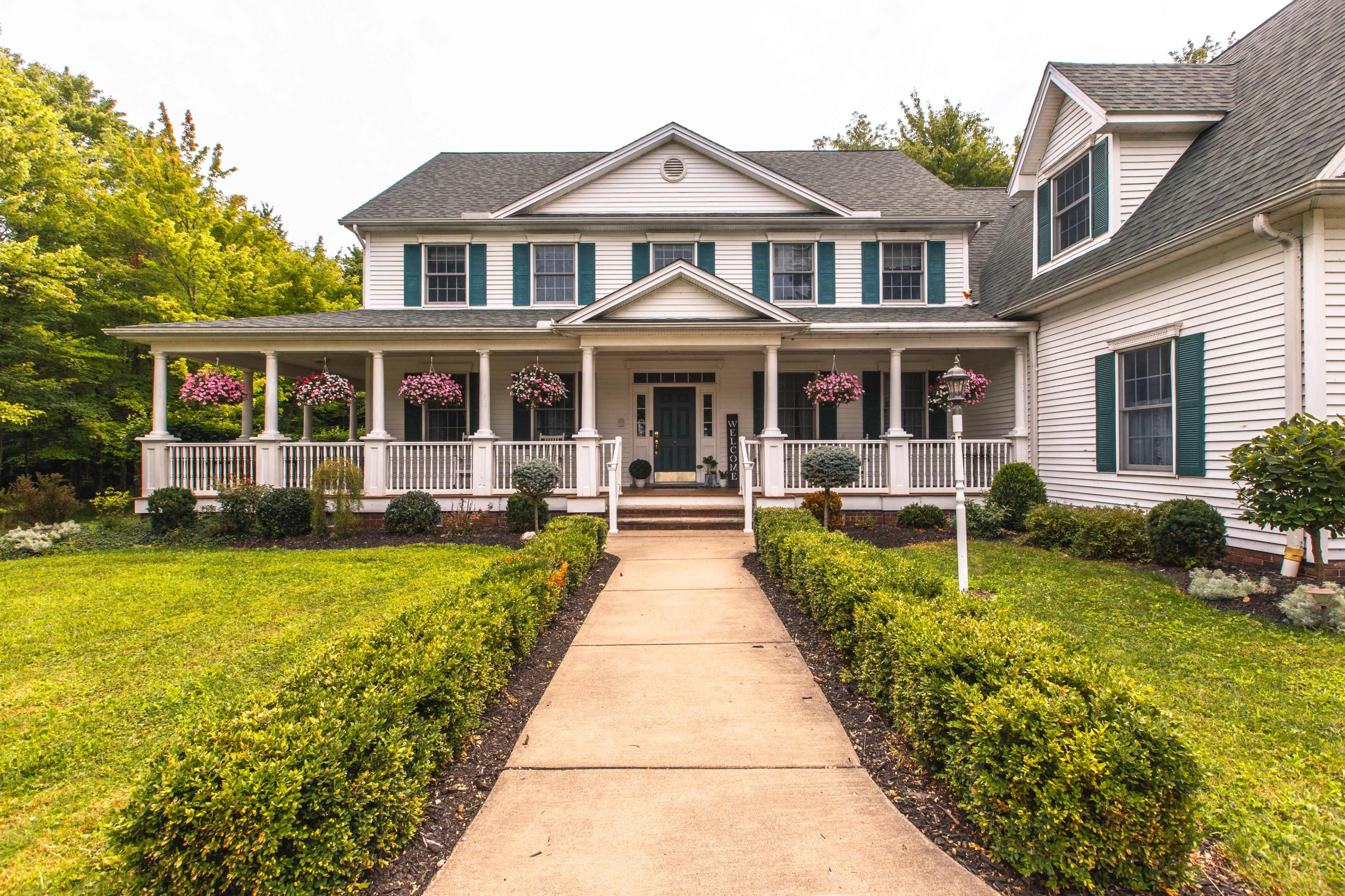 A large, two-story house with a front porch and hanging flower baskets is surrounded by a neatly landscaped yard and pathway.