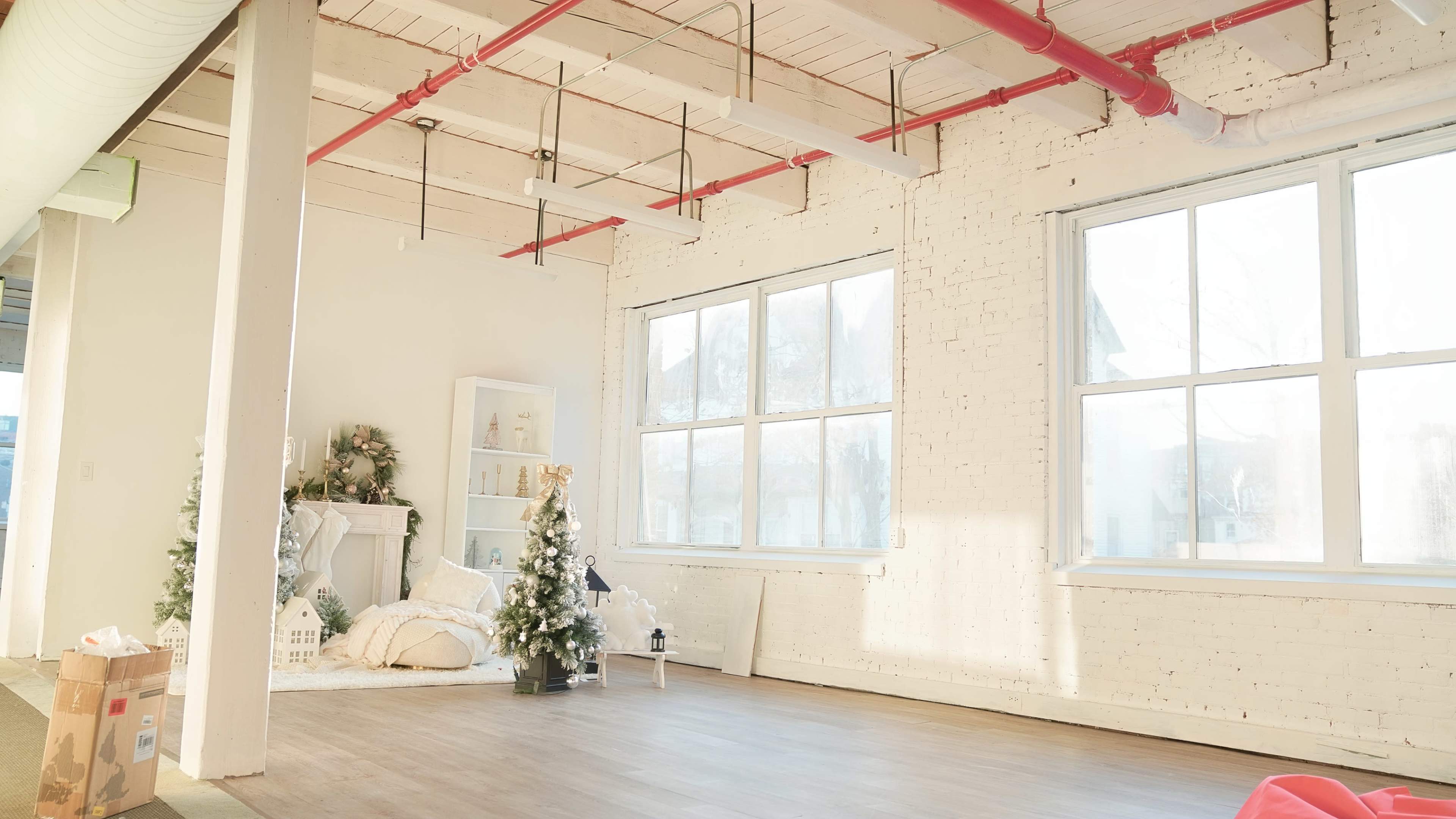 A bright, spacious room with large windows features a small Christmas tree, decorative items on a shelf, and minimal furniture against a white brick wall.