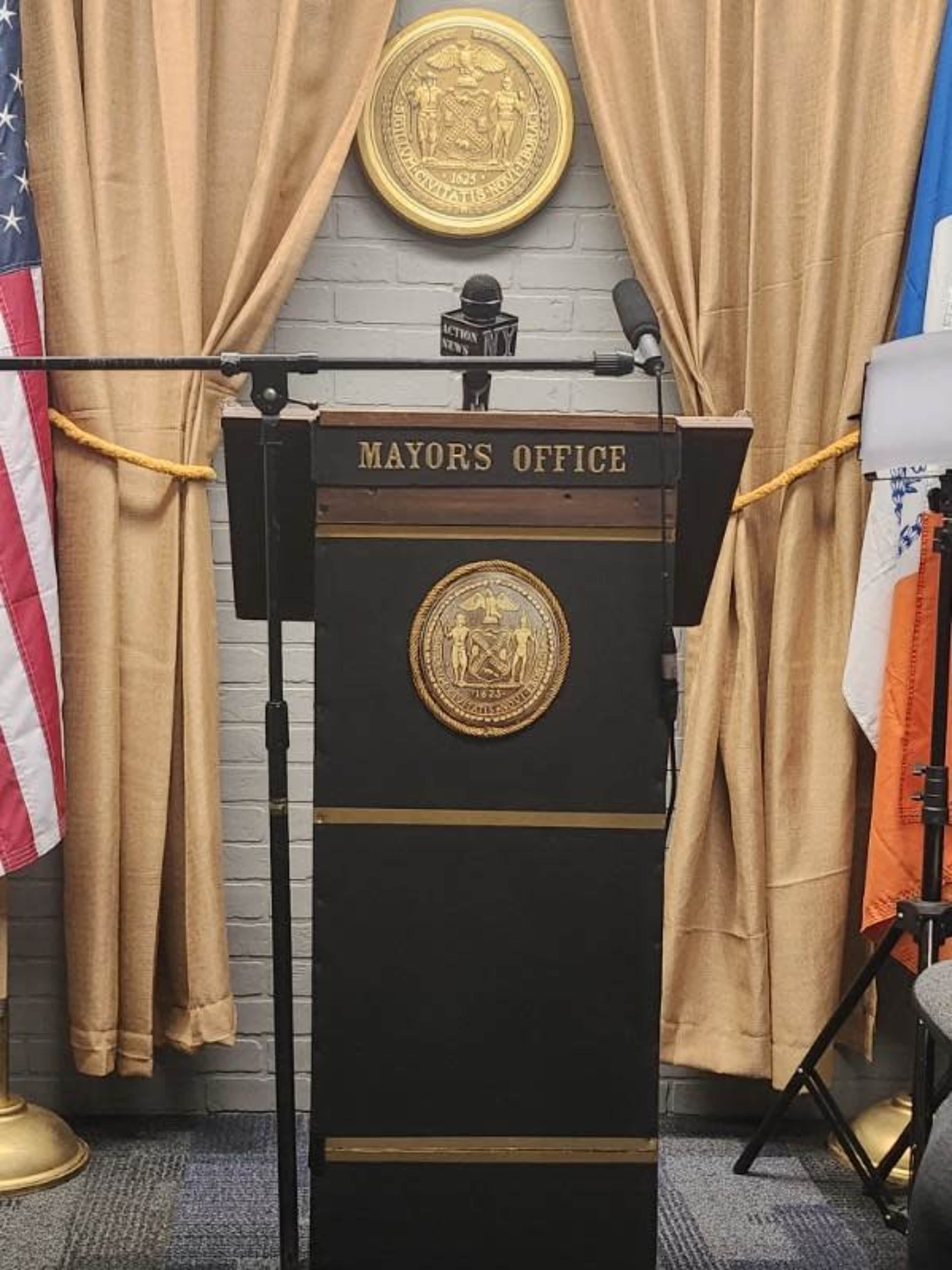 A podium labeled "MAYOR'S OFFICE" with a microphone and a backdrop featuring curtains, an American flag, and city emblems.