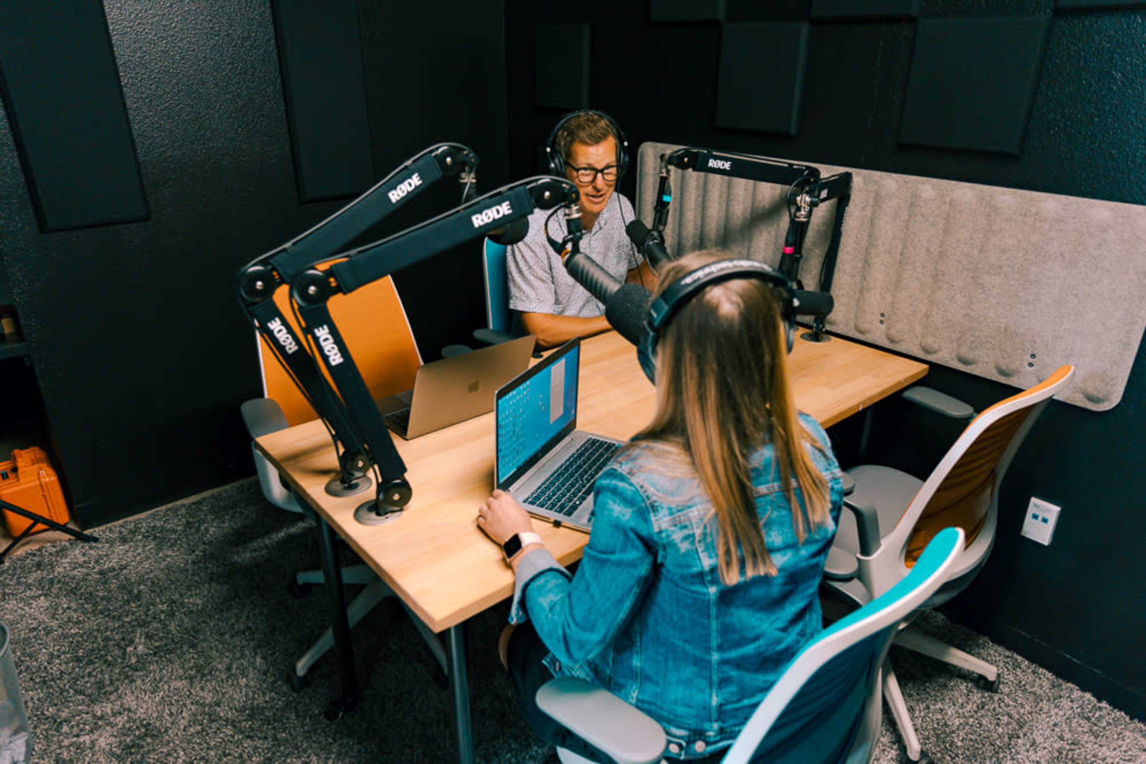 Two people sit at a table in a recording studio equipped with microphones and laptops, engaged in conversation.