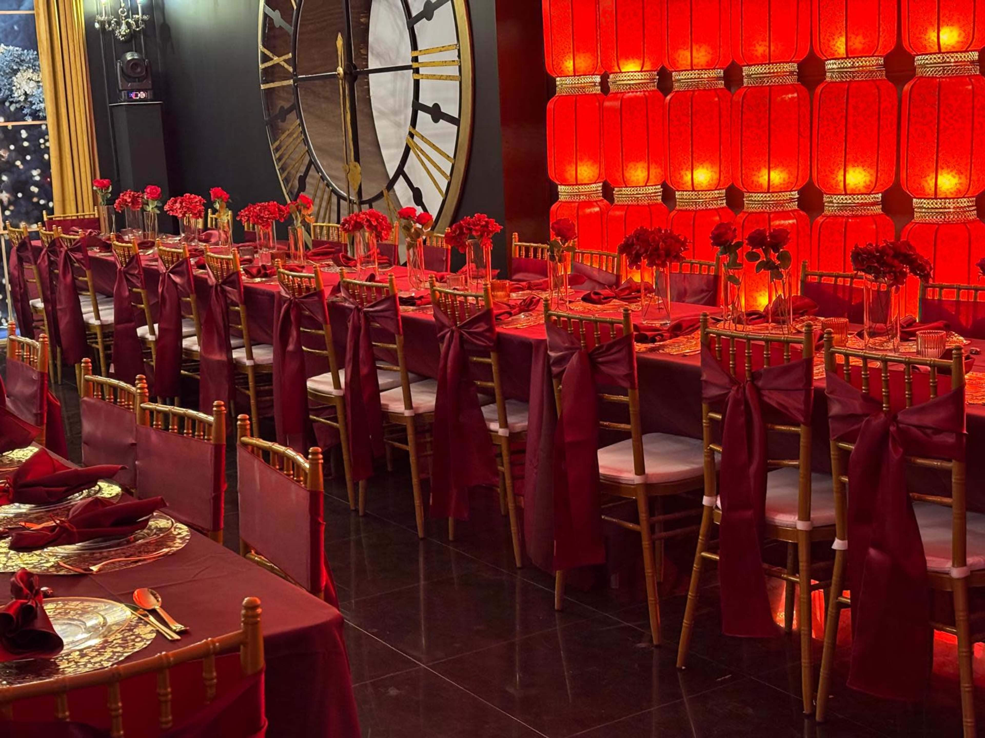 The image shows a banquet hall arranged with long tables adorned with maroon tablecloths, golden chairs, and centerpieces of red flowers, under a backdrop of illuminated red panels and a large clock.