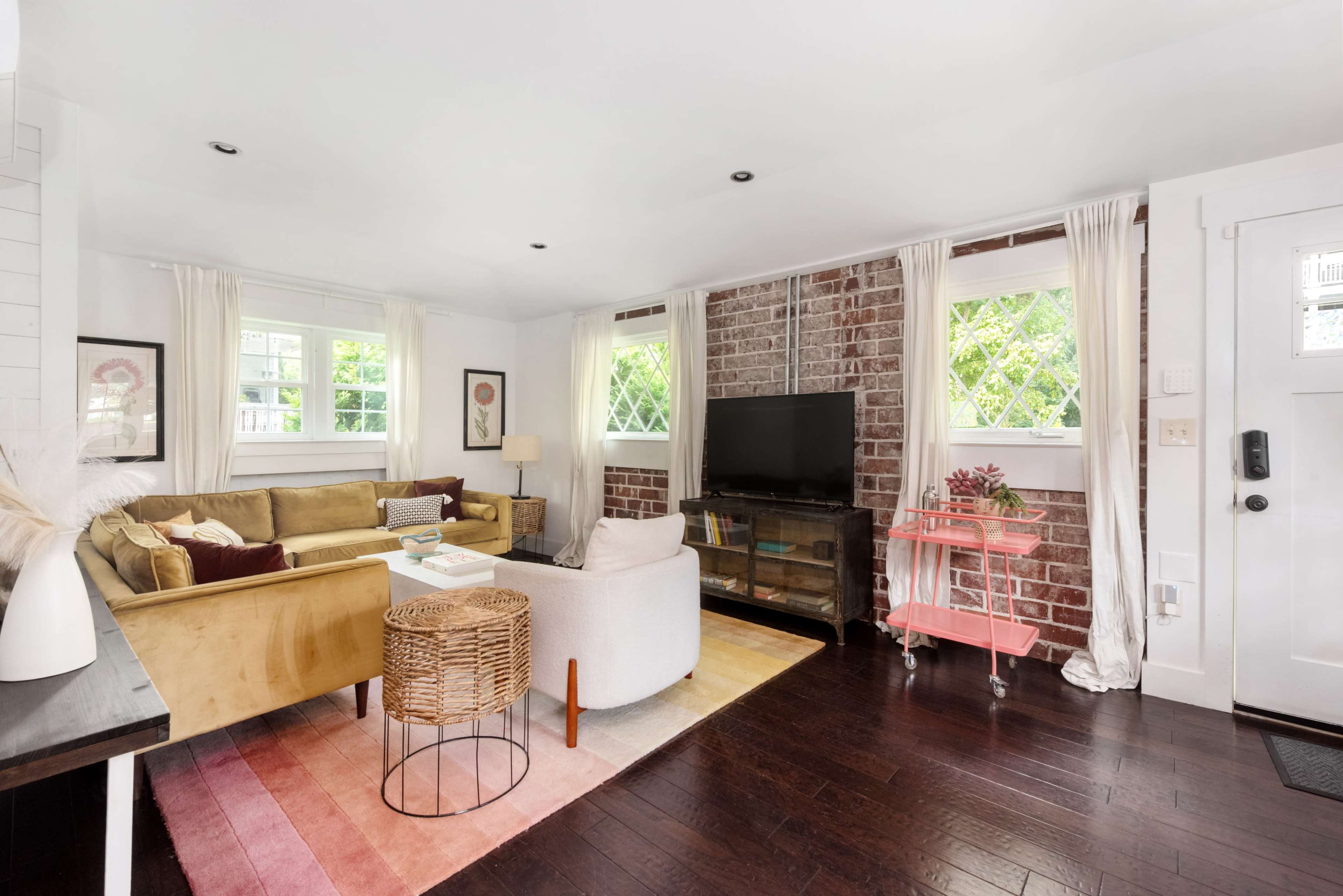 The living room features a sectional sofa, a television against a brick wall, and large windows with curtains, all set on dark hardwood flooring.