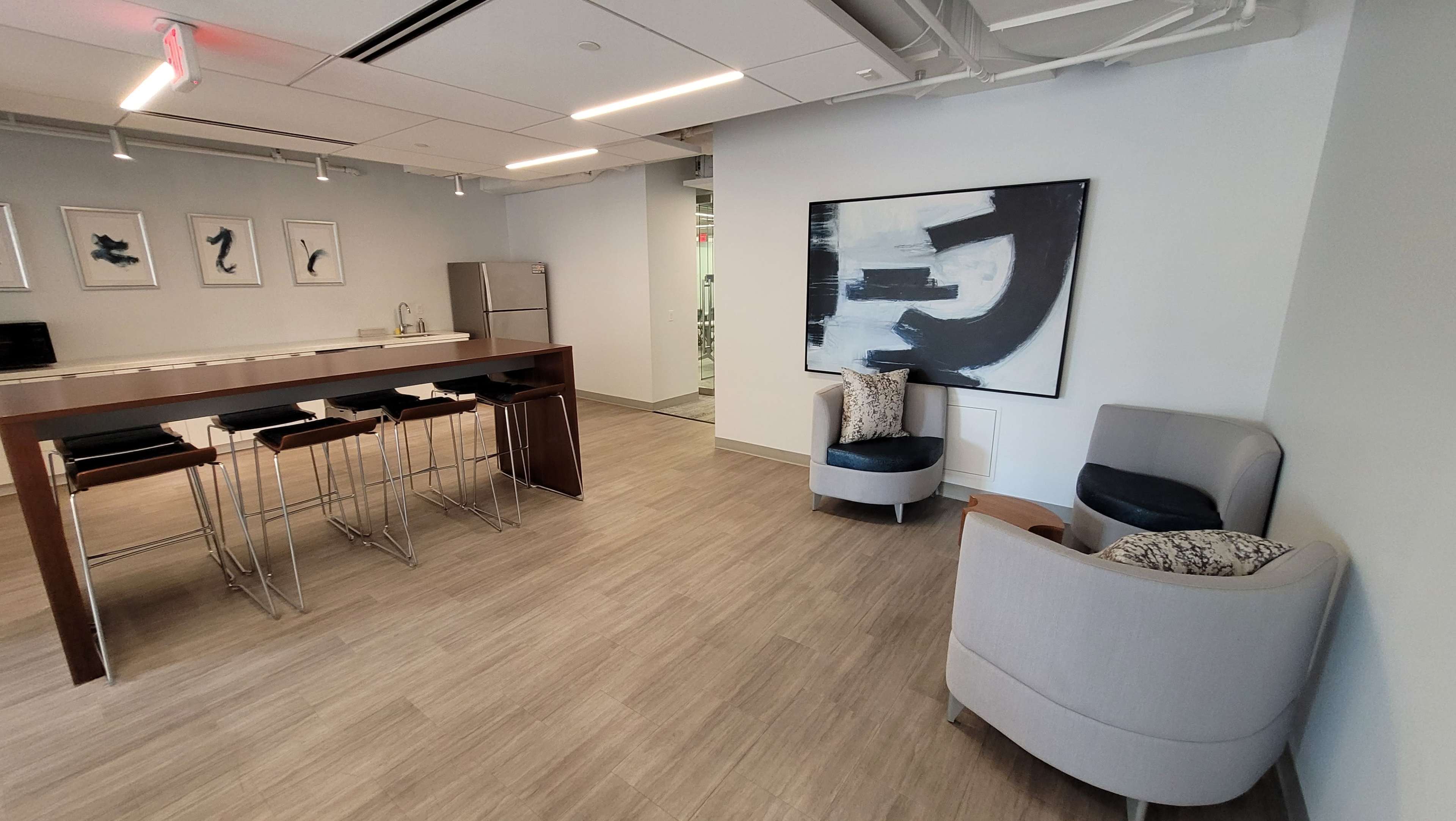 The image depicts a modern office lounge area featuring a kitchenette with bar stools, a seating area with two armchairs, and a large abstract artwork on the wall.