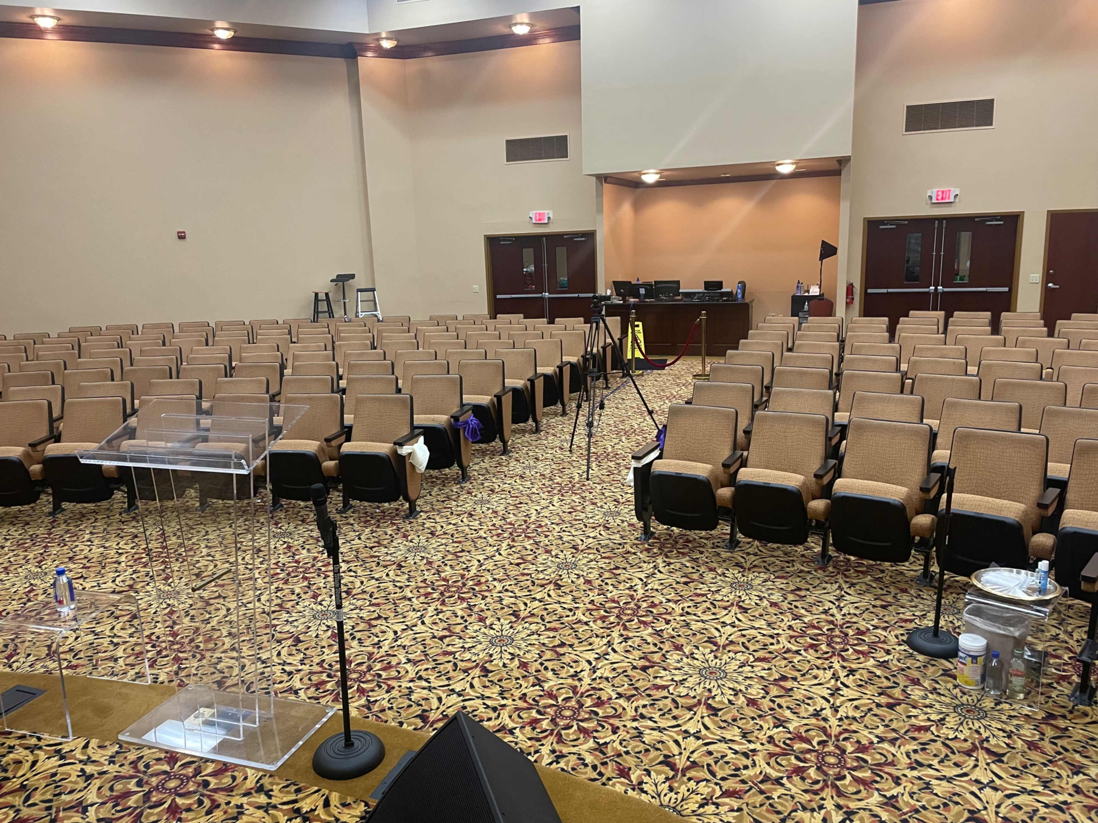 The image shows a spacious auditorium with rows of empty seats, a clear podium at the front, and a setup for filming or broadcasting, including tripods and equipment.