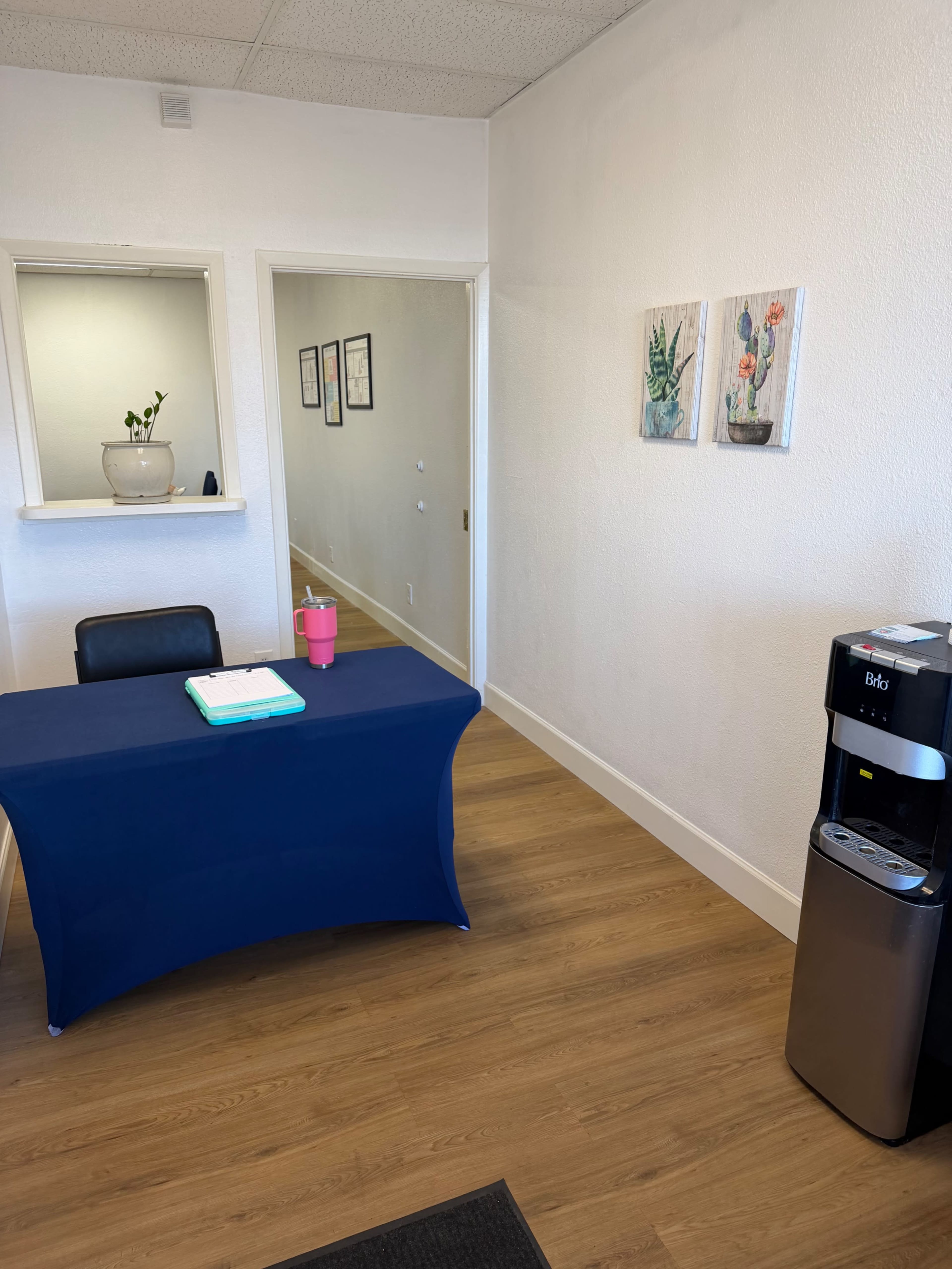 The image shows a bright, empty office space featuring a desk with a blue tablecloth, a water cooler, and wall art depicting plants.