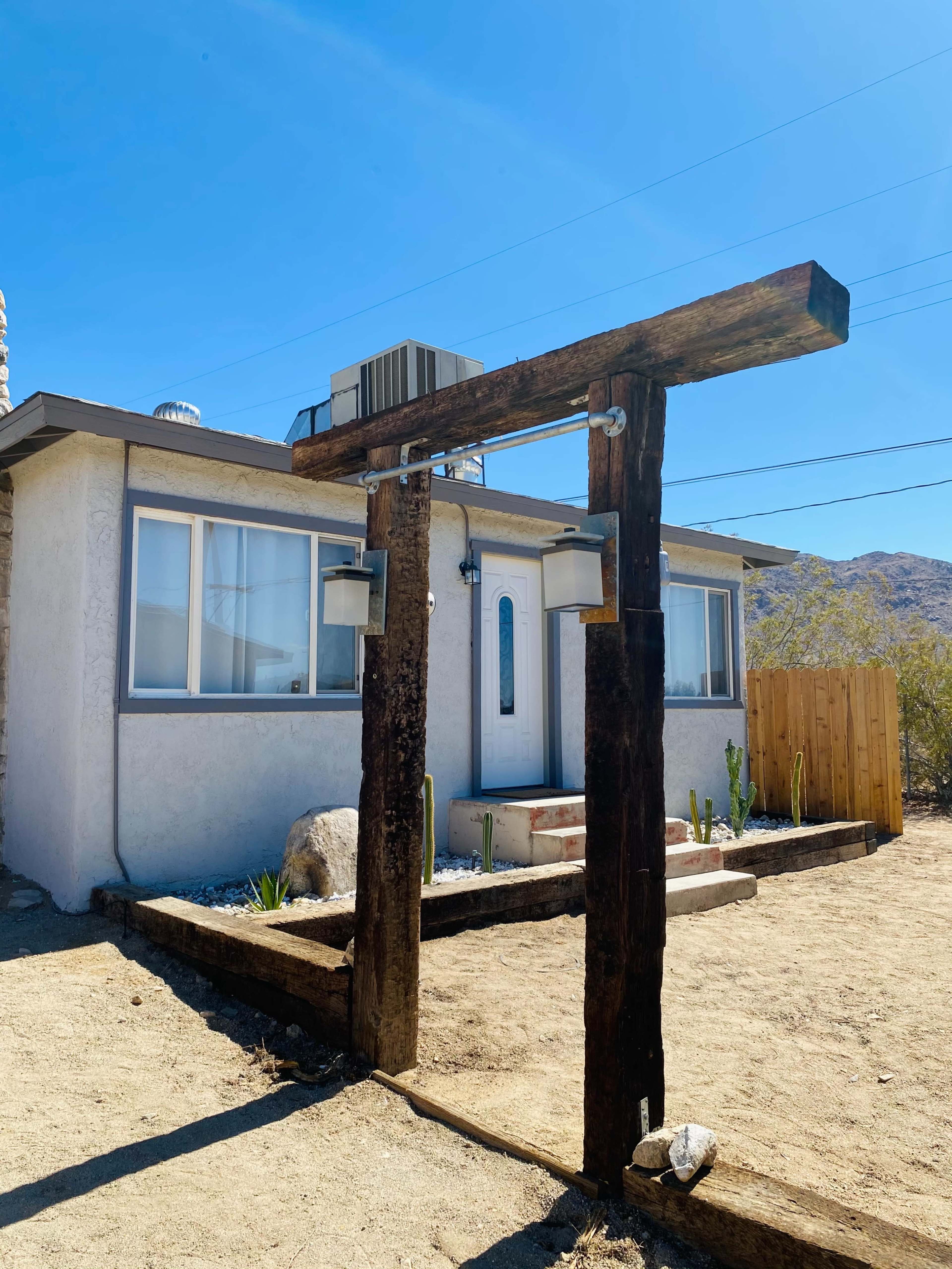 A rustic wooden arch with lanterns frames the entrance to a single-story house surrounded by gravel and desert landscape.