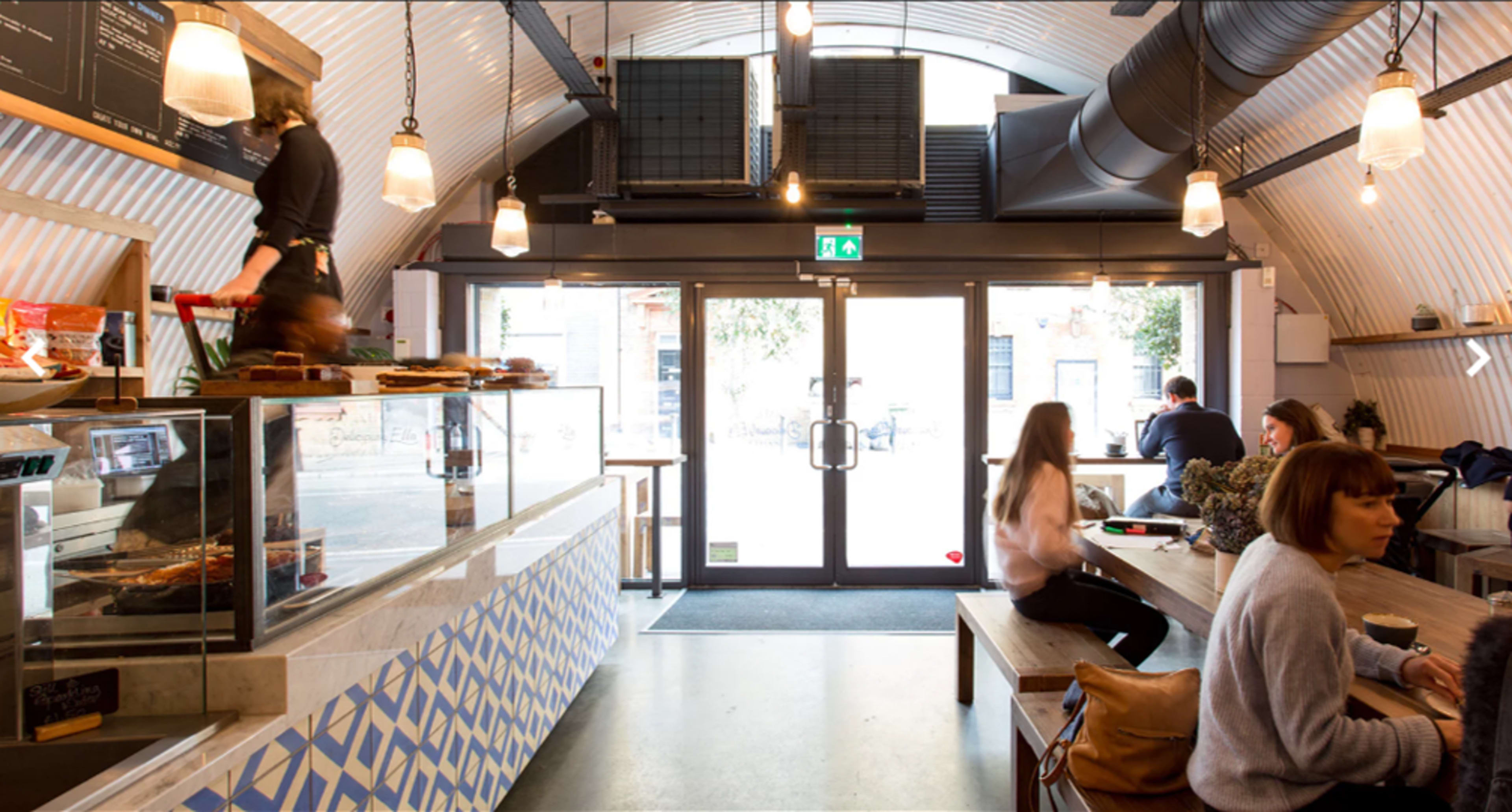 The interior of a café features a service counter with pastries, wooden benches, and large windows providing natural light.