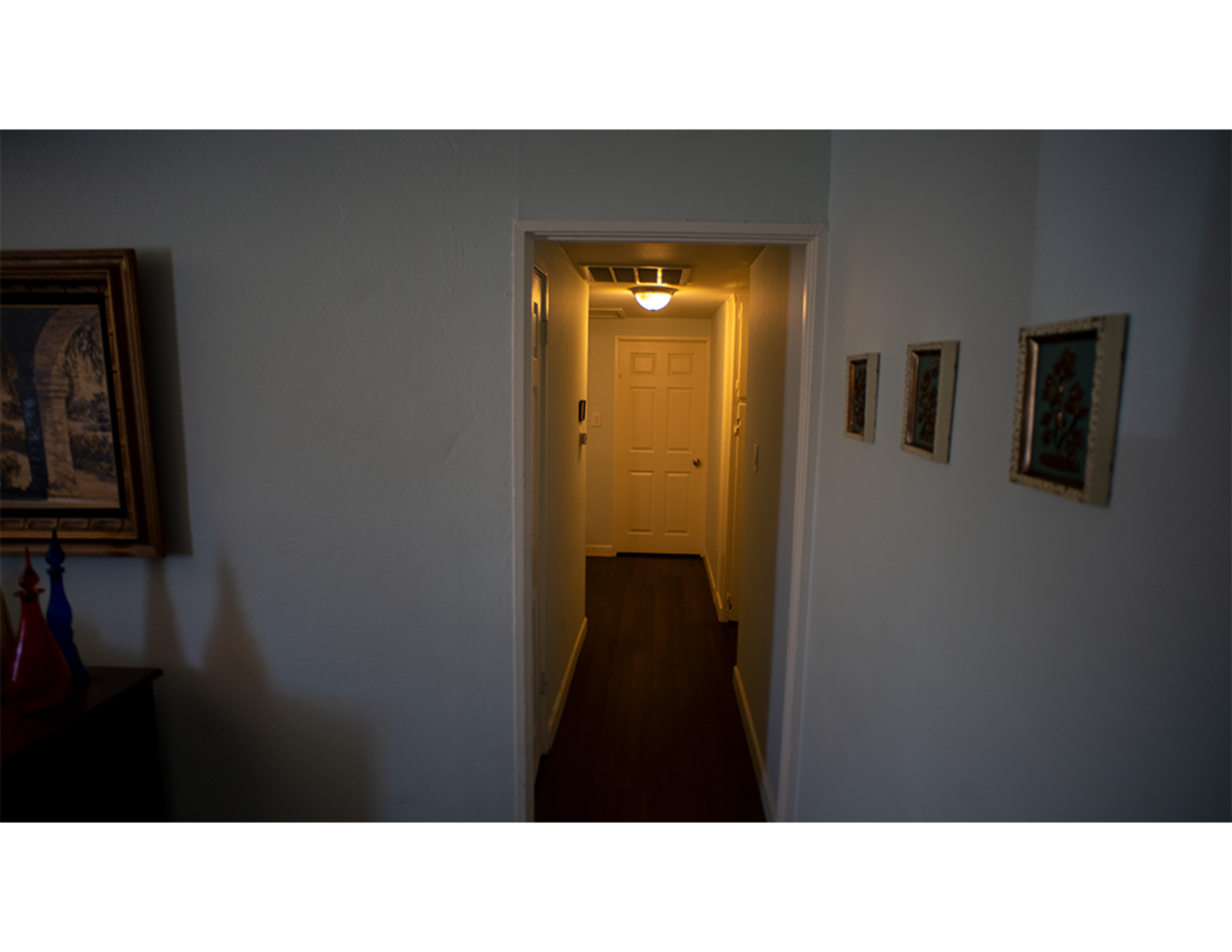 A well-lit hallway leads to a closed door at the end, framed by light-colored walls and adorned with small decorative pictures.