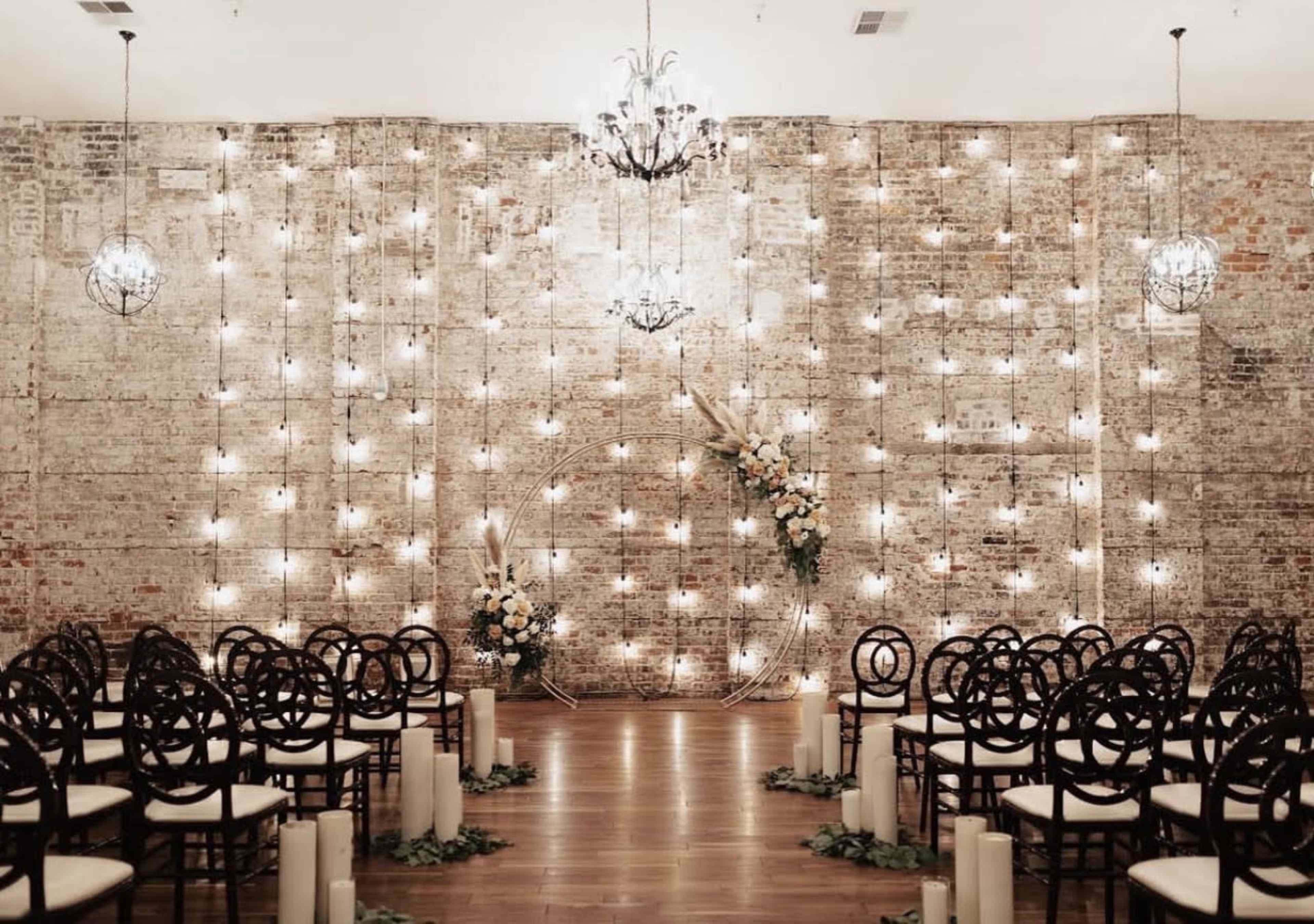 A decorated venue features a brick wall adorned with lights and floral arrangements, with black chairs arranged in rows facing a circular floral arch.