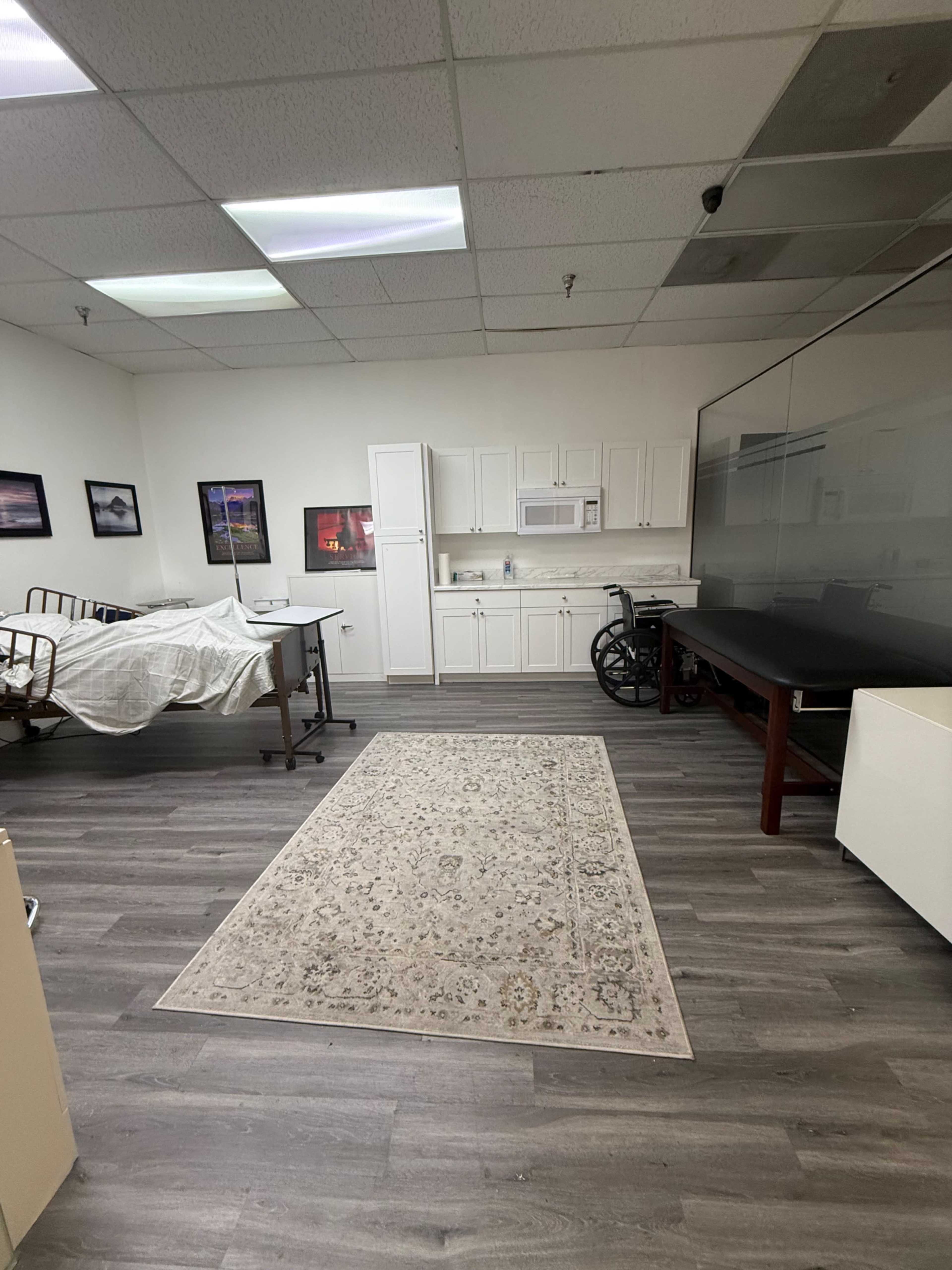 The image shows a spacious room with a bed partially covered by a sheet, a table, a wheelchair, and a kitchenette area with white cabinets and a microwave.