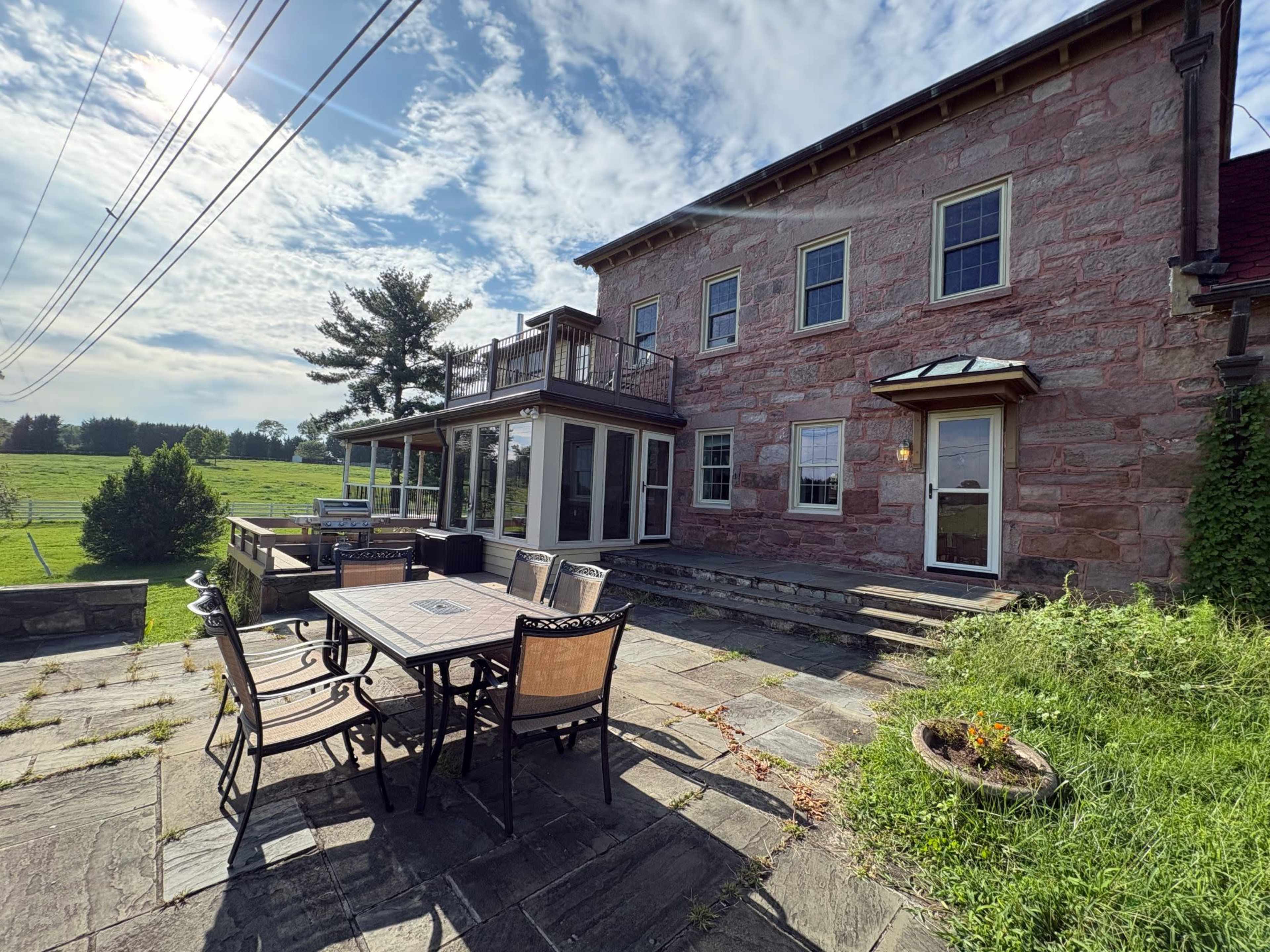 The image shows a spacious stone patio with outdoor dining furniture in front of a large, two-story building surrounded by green fields and trees under a cloudy sky.