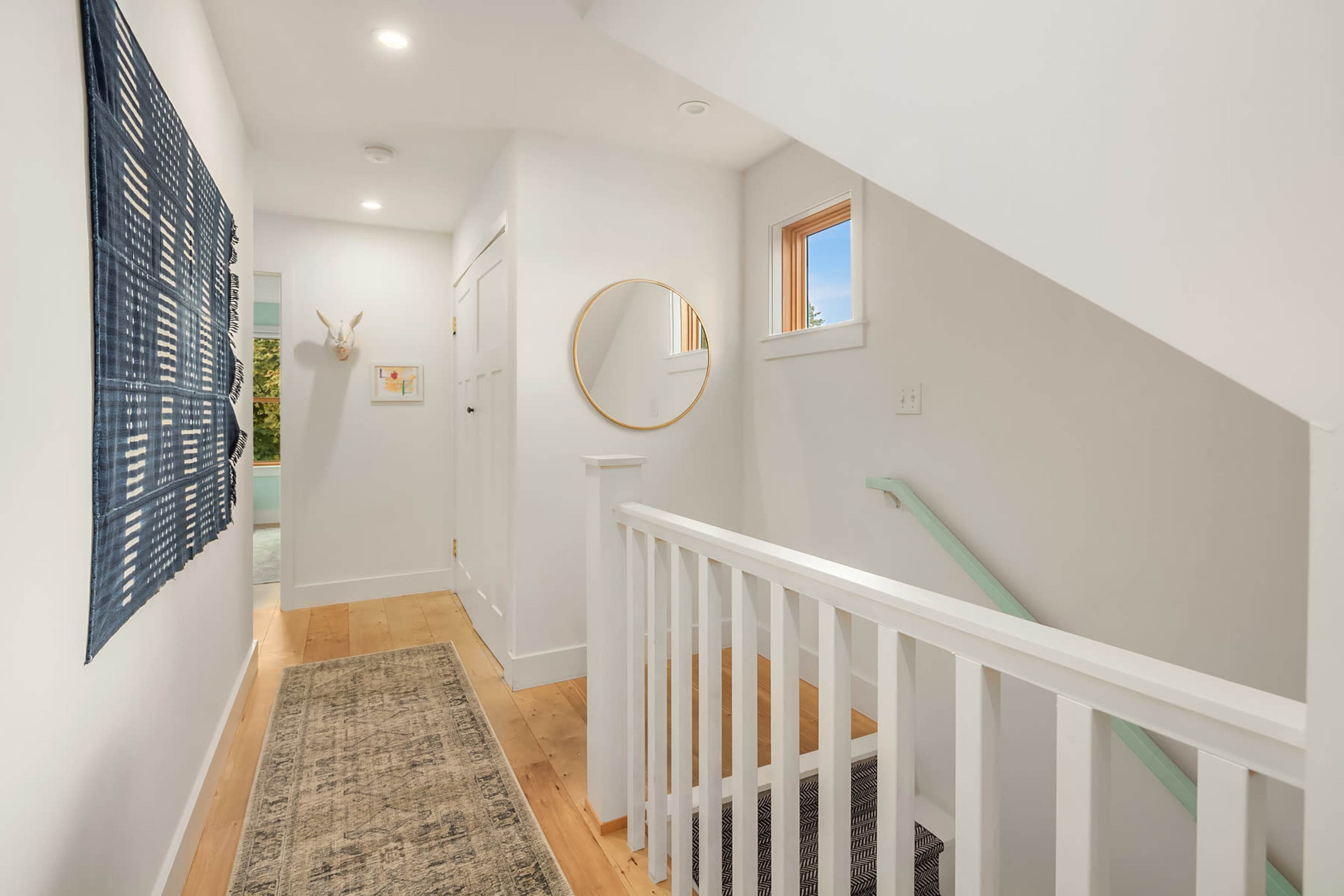 The image shows a bright hallway with a staircase, adorned with a wall mirror, a decorative wall hanging, and a rug along the wooden floor.
