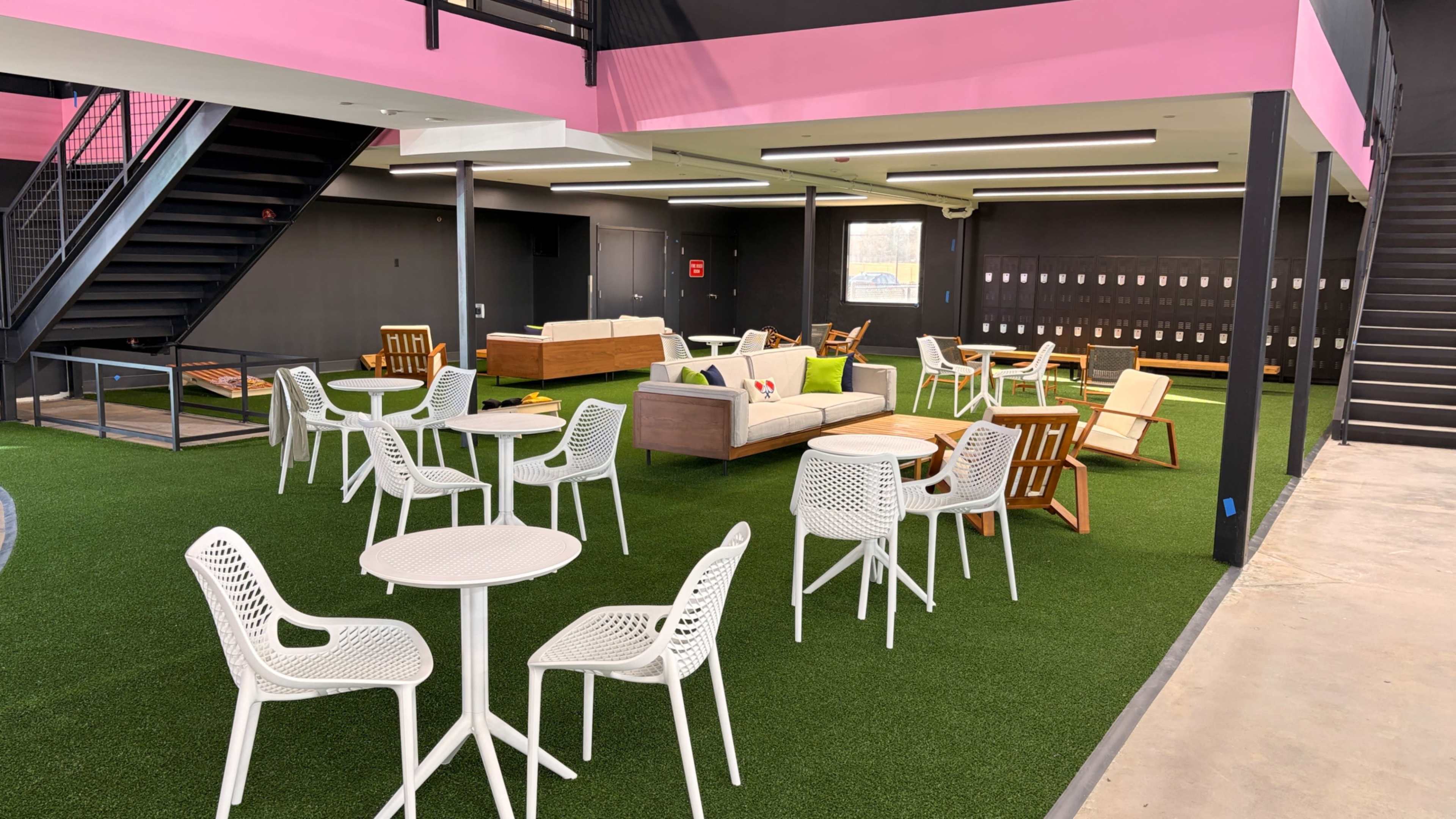 The image shows a modern indoor lounge area with artificial grass, featuring white tables and chairs, colorful sofas, and a staircase in the background.
