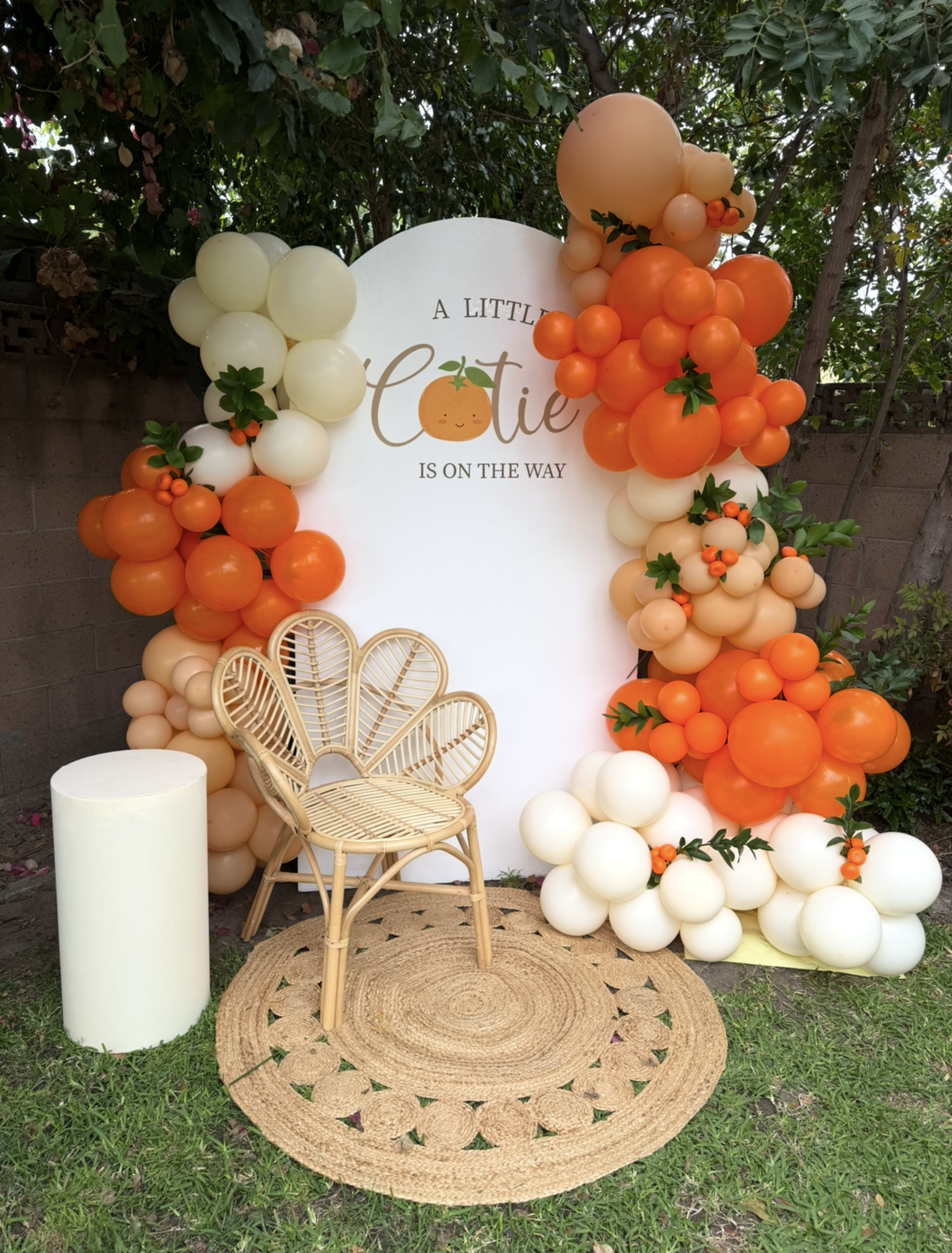 The image features a festive backdrop decorated with orange and cream balloons arranged around a circular sign that reads "A Little Cutie Is On The Way," alongside a woven chair and a round rug on the grass.