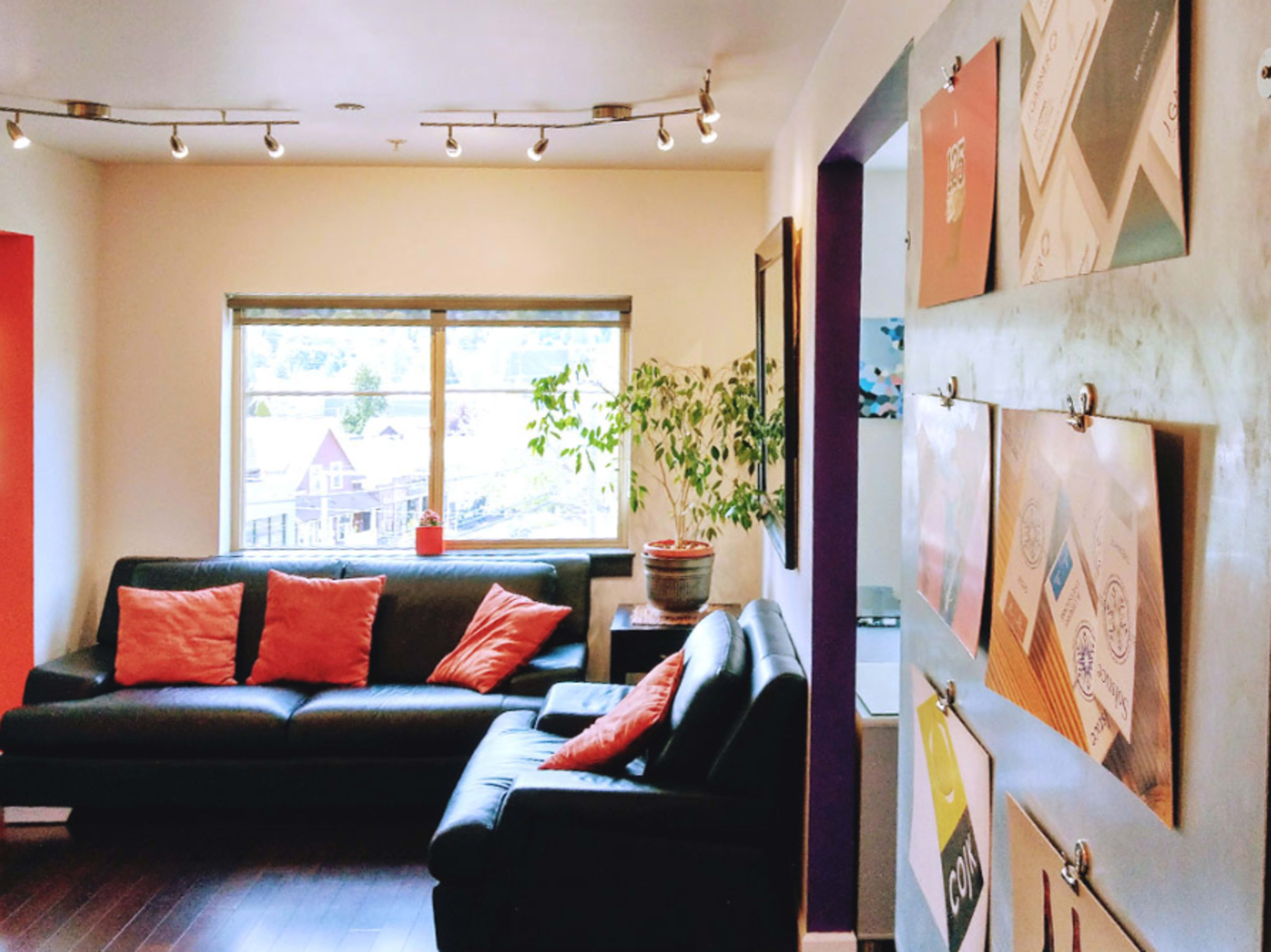 A living room featuring two black sofas, orange pillows, a large window with a view, and a wall displaying various colored cards or artwork.