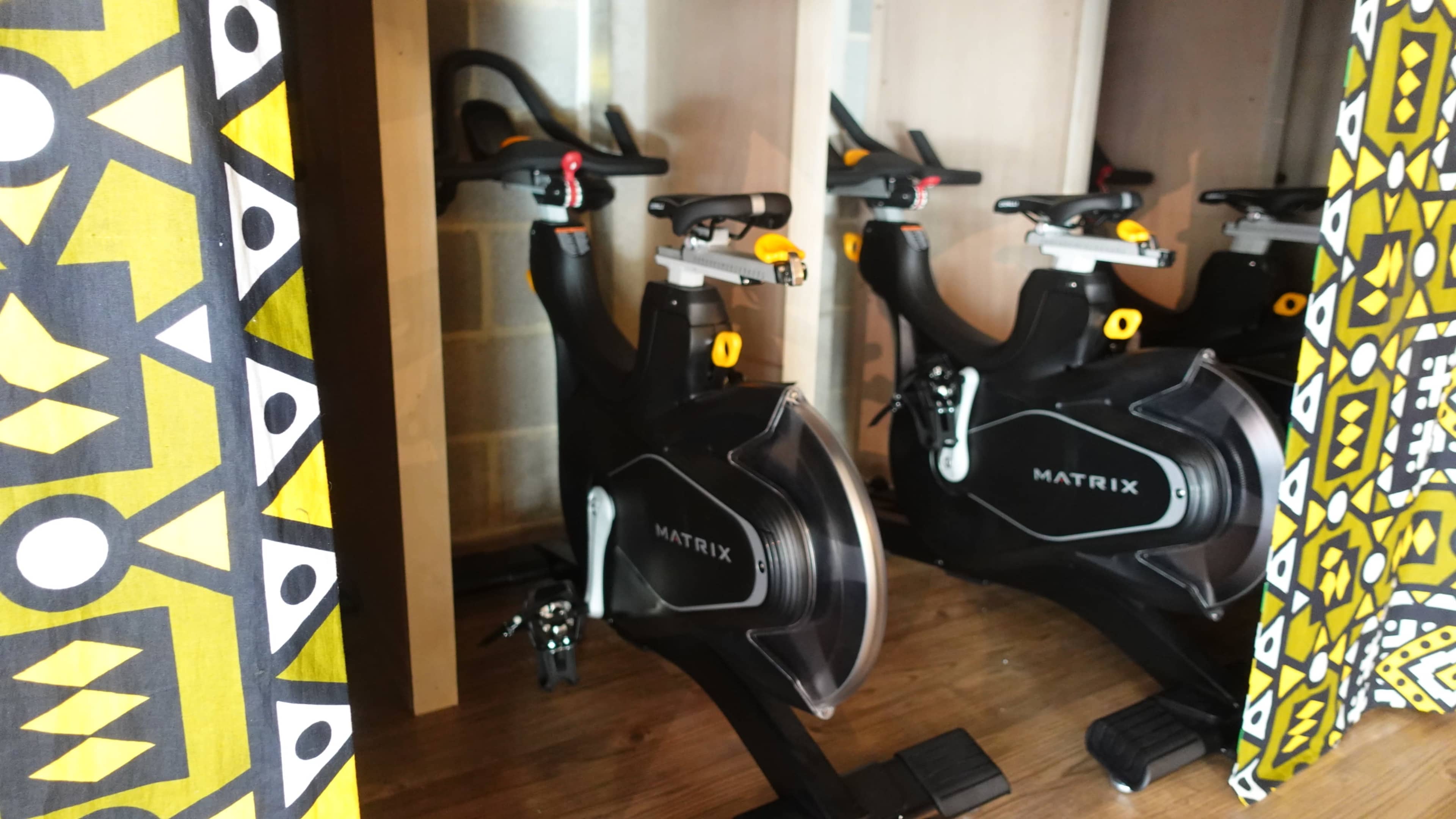 The image features two exercise bikes positioned within a fitness studio, partially obscured by patterned curtains.