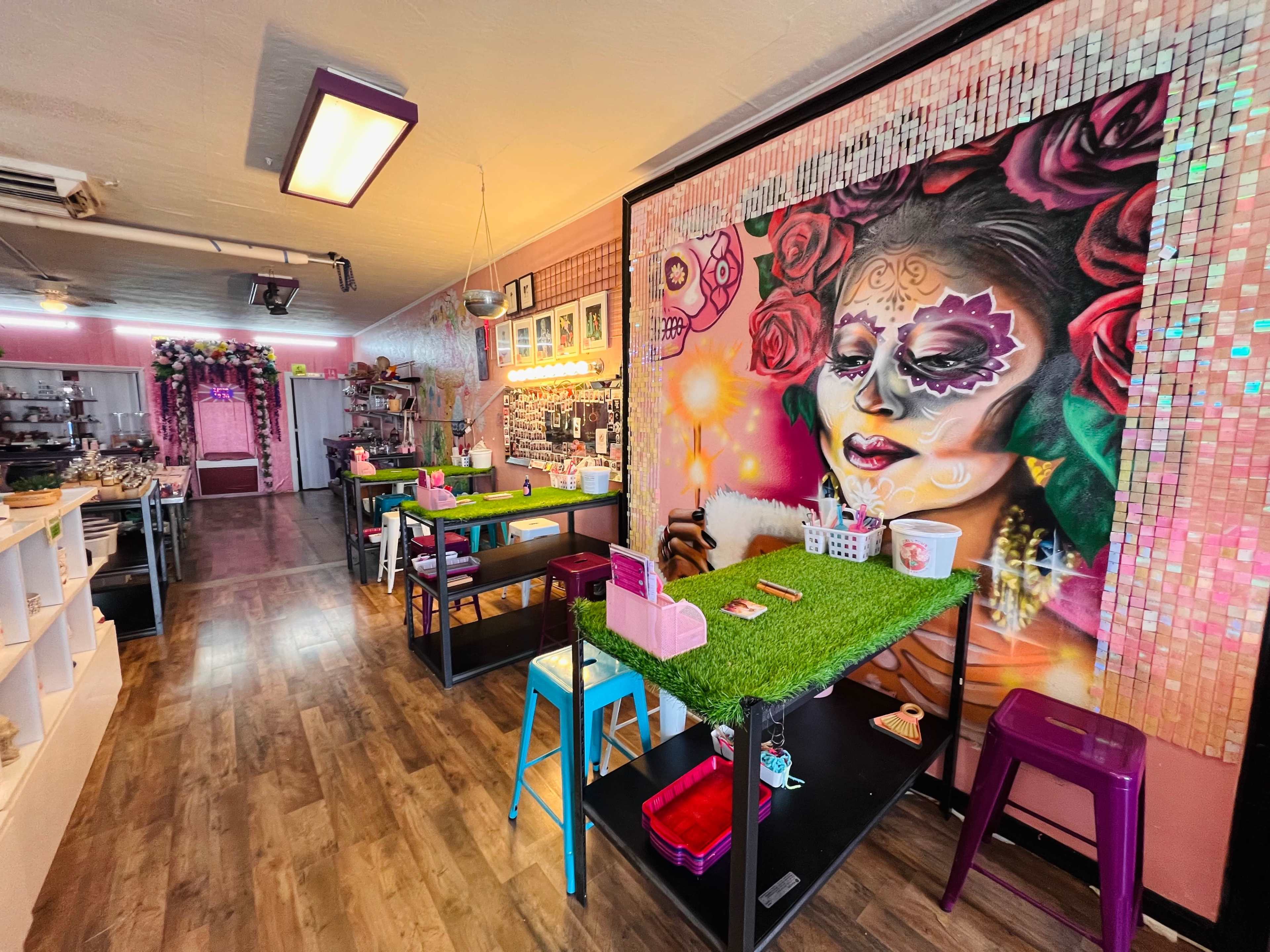 The interior of a colorful studio features tables with green turf tops, vibrant artwork on the wall, and shelves displaying various craft supplies.