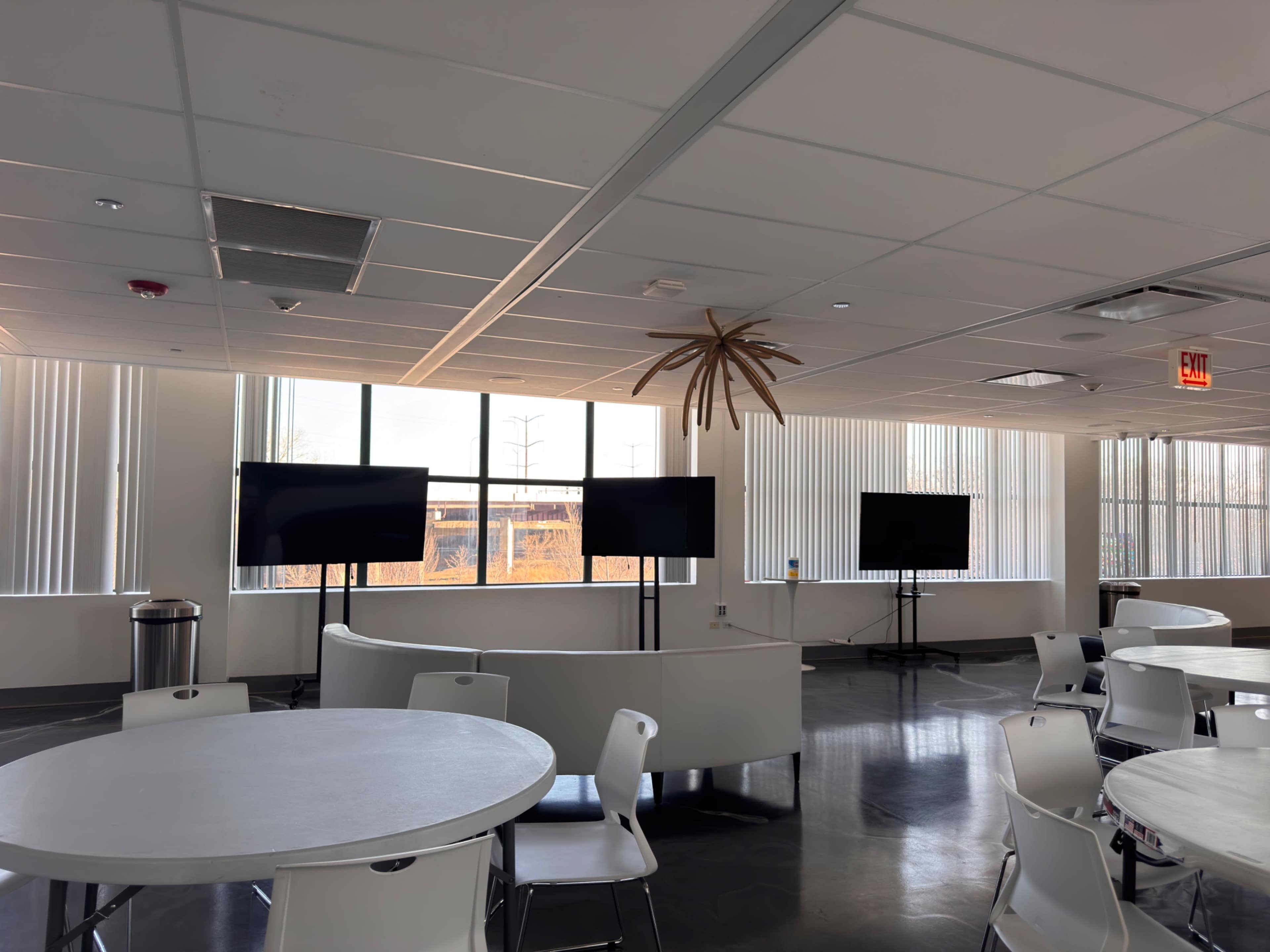 The image shows a modern, brightly lit room with several round tables, white chairs, and multiple flat-screen televisions mounted on stands near large windows.