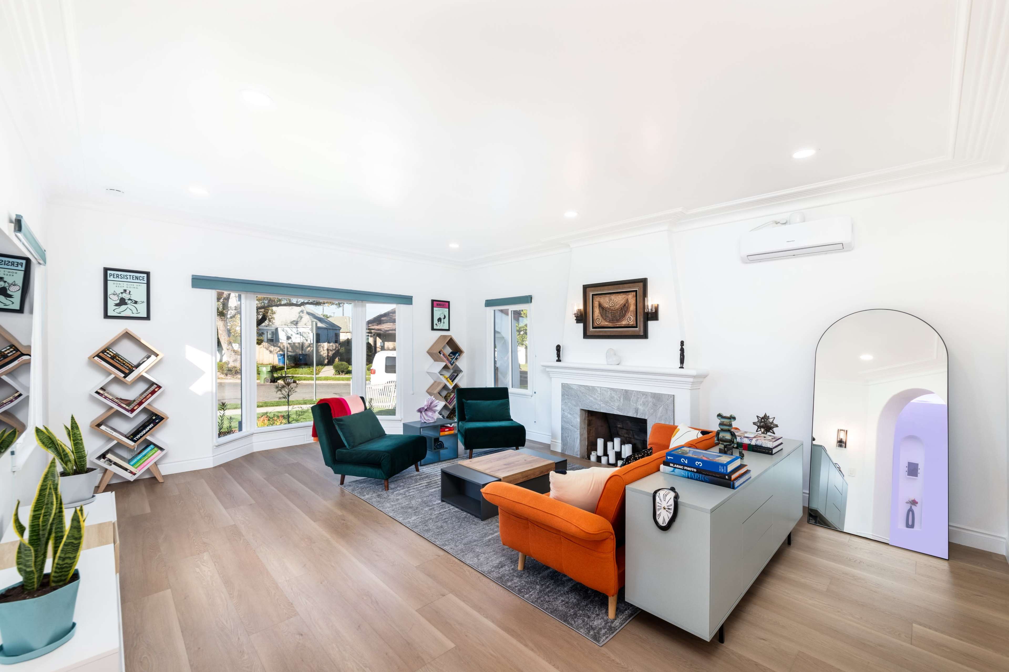 A modern living room features a fireplace, colorful seating, bookshelves, and large windows providing natural light.