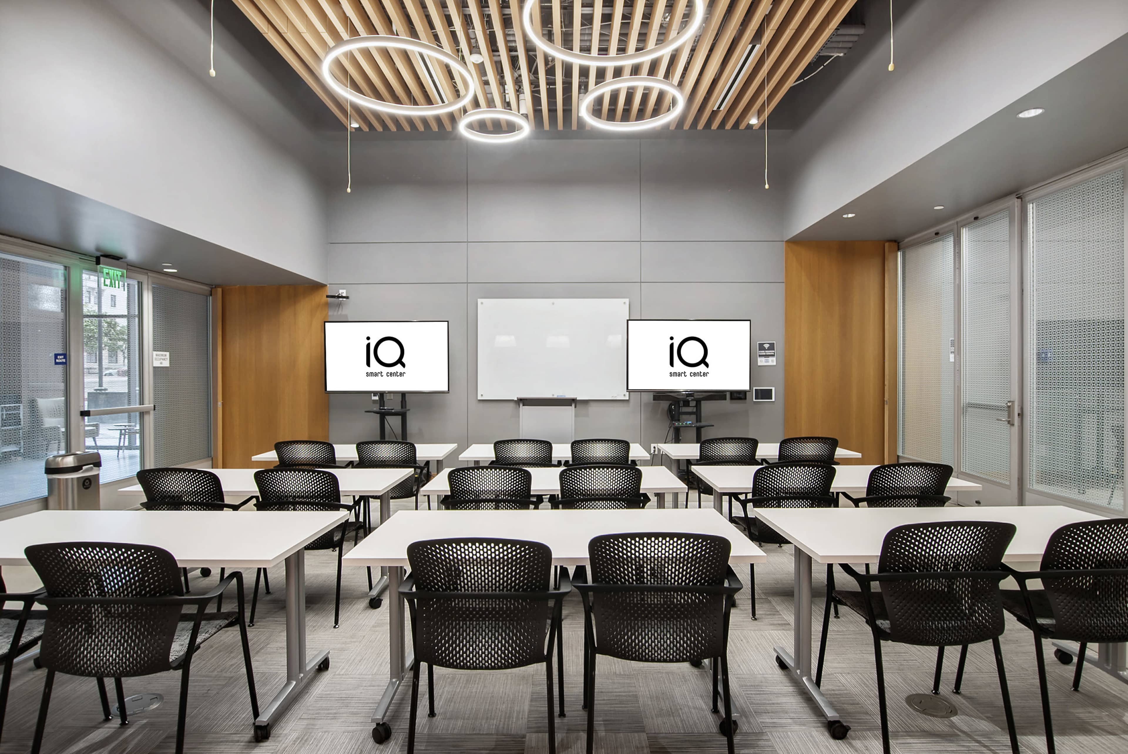 A modern classroom features white tables arranged in rows, black chairs, and two screens displaying a logo on the front wall.