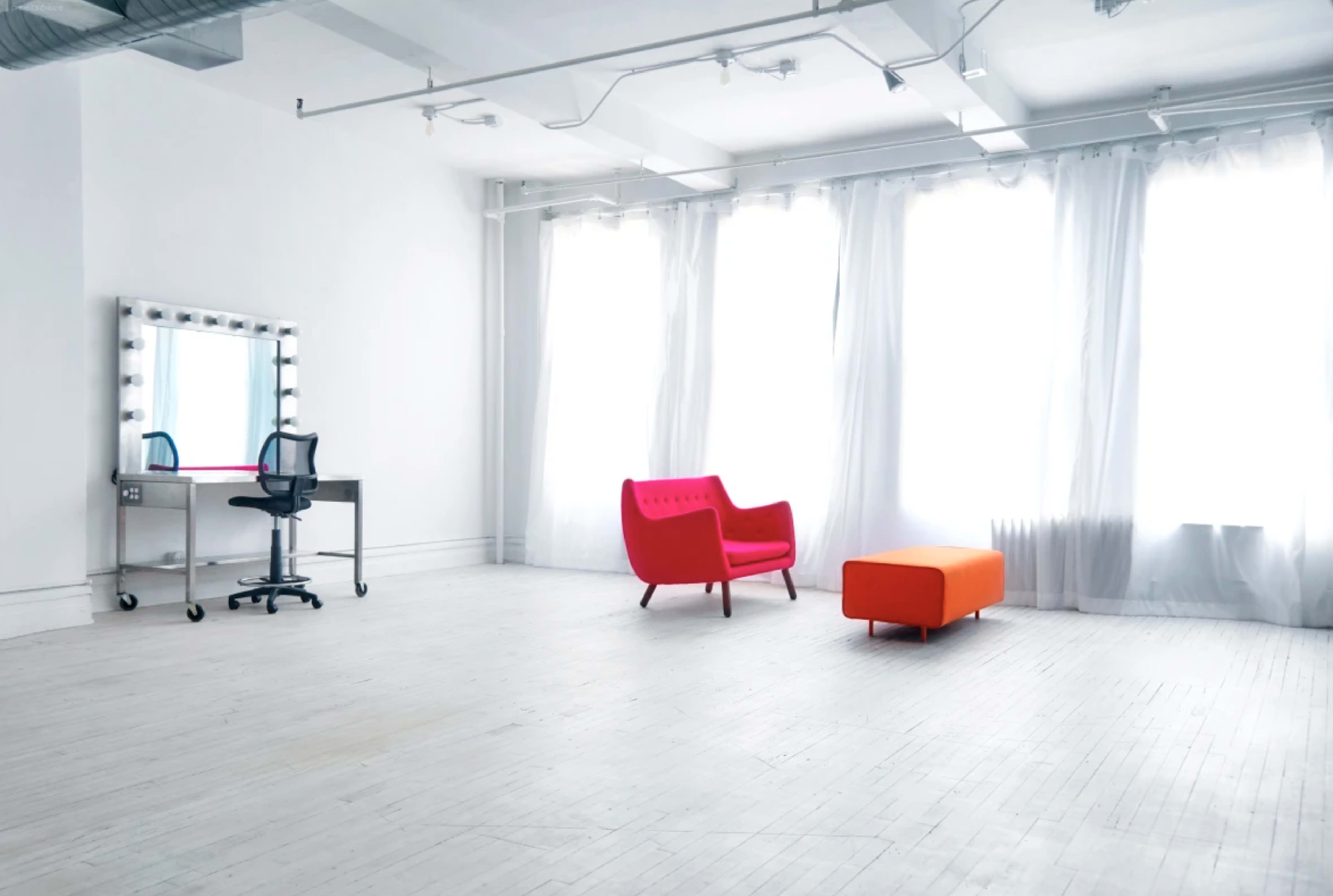 A bright, minimalist interior featuring a vanity table with a chair, a red sofa, and an orange ottoman near large windows with sheer curtains.