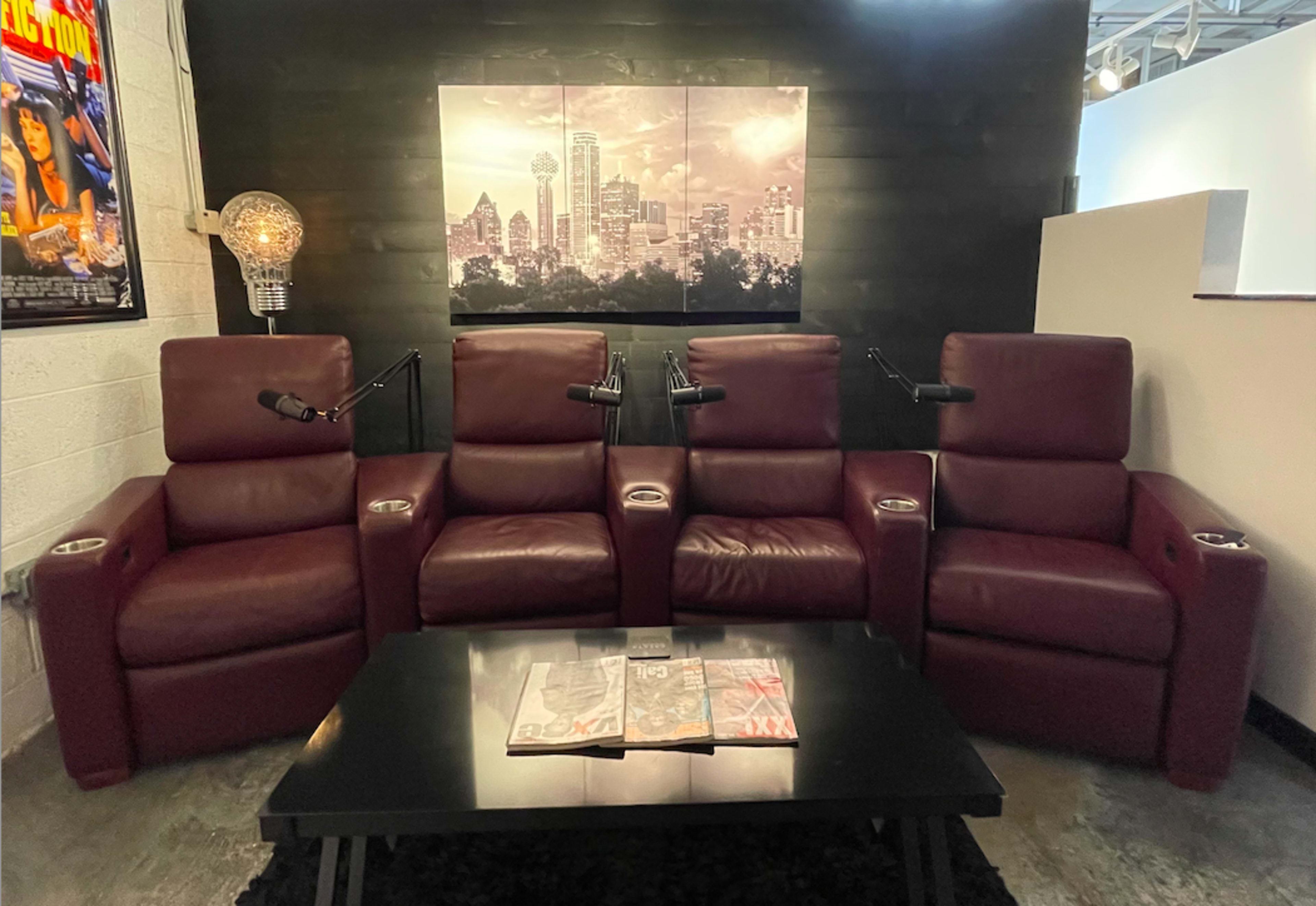 A row of three burgundy recliners with adjustable reading lights and a low black table featuring magazines in front of them, set against a wall with a cityscape artwork.