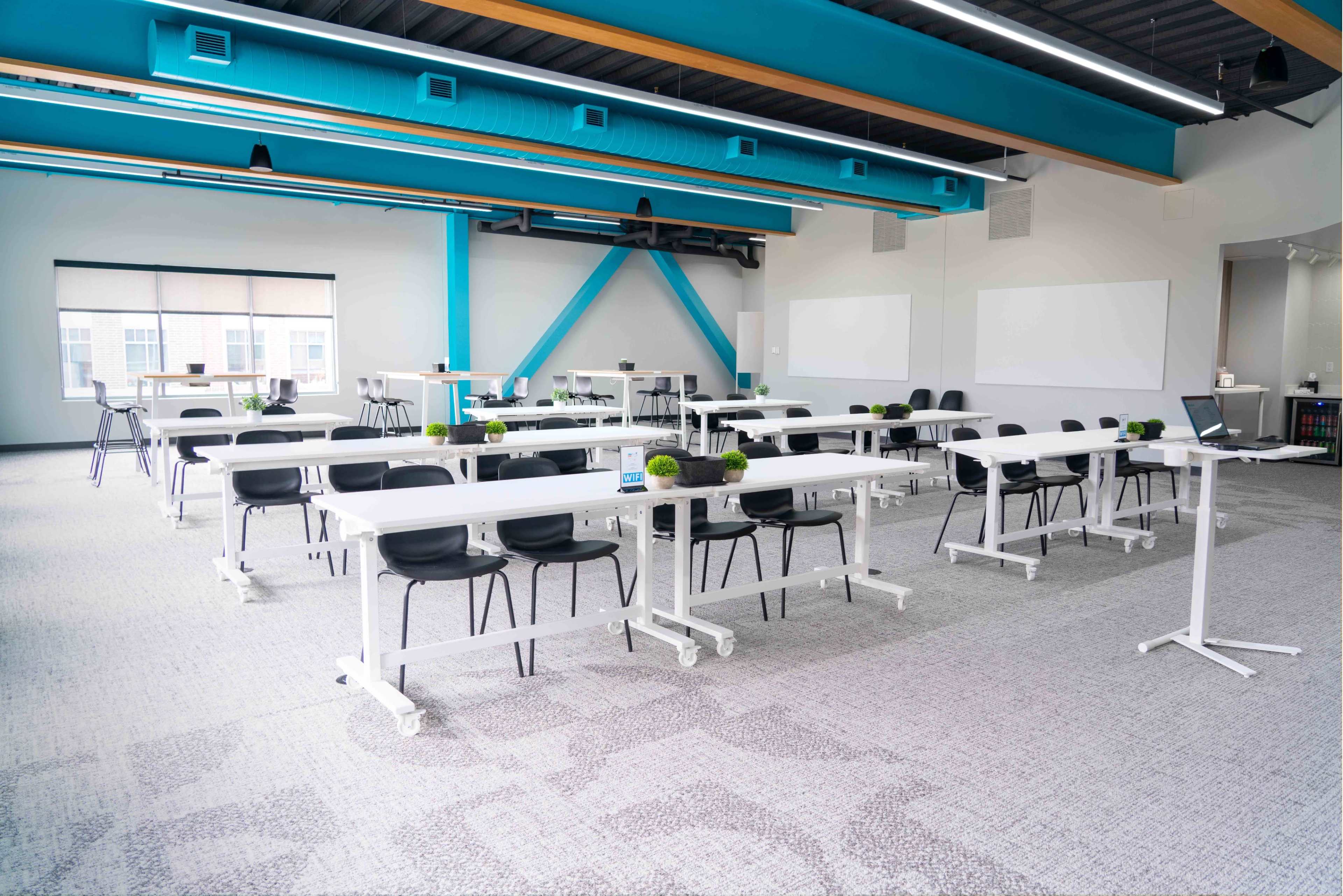 A modern conference room features multiple white tables and chairs arranged in a spacious, well-lit layout with green decor accents.